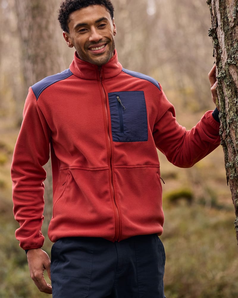 A man standing in a forest wearing a red fleece with blue patches.