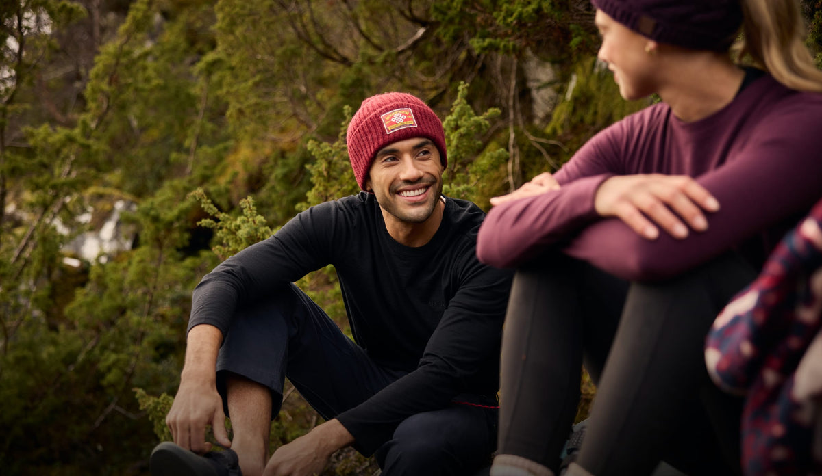 Man wearing a red knit beanie with a visible Sherpa Adventure Gear logo, sitting and talking in a wooded setting.