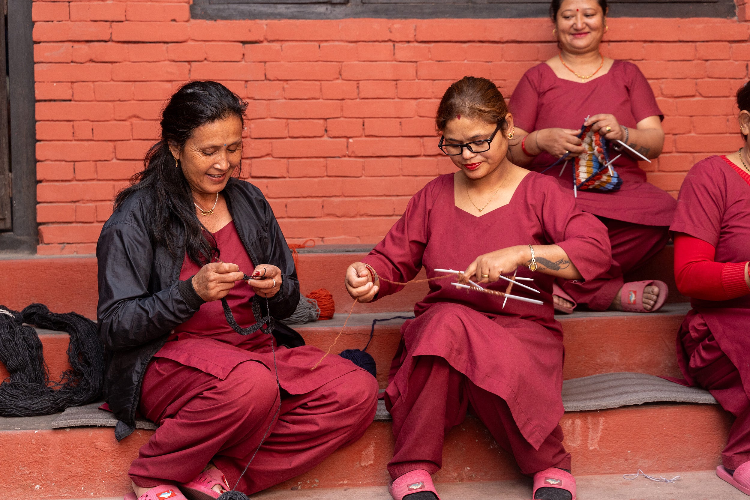 A group of women in Nepal sitting on a set of steps. They are hard at work  knitting hat and knitted accessories by hand.