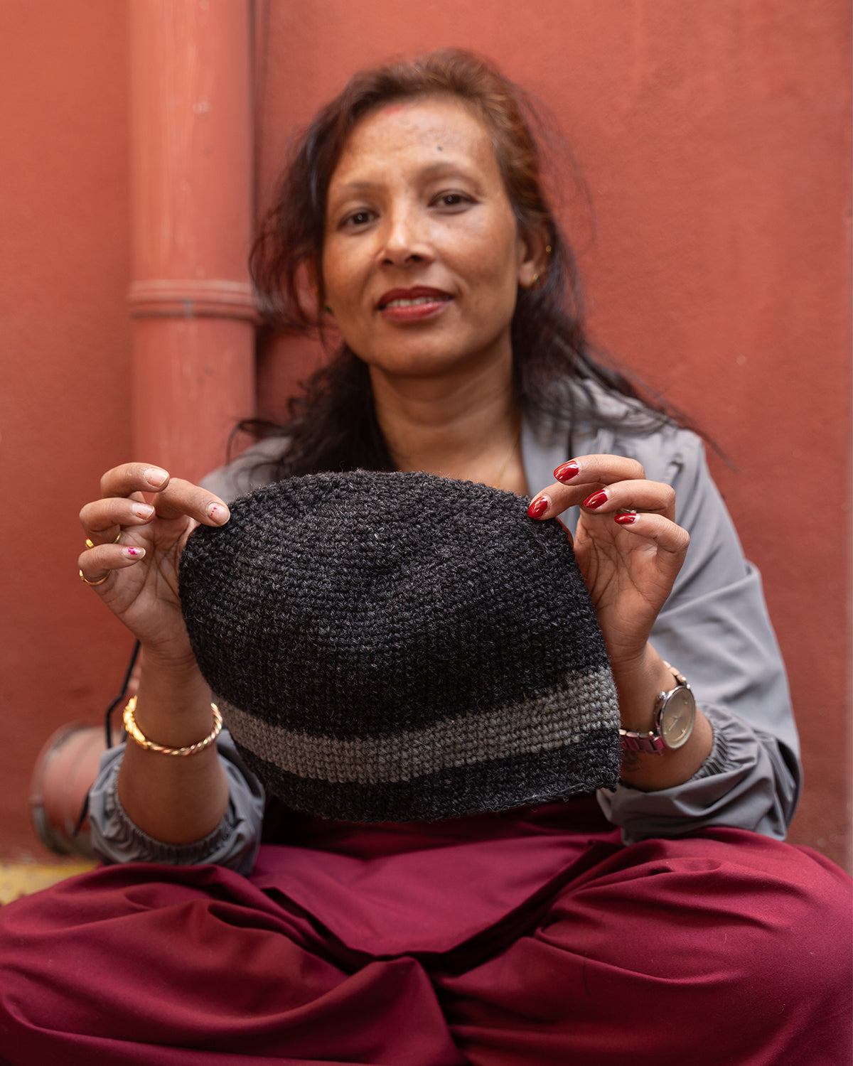 A woman in Nepal smiling at the camera whilst holding up a knitted hat. She is wearing a blue over-shirt and a red set of trousers.