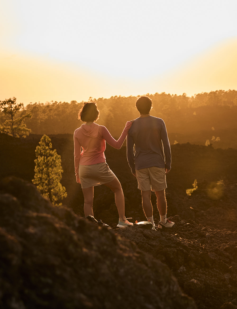A man and a woman standing on a rocky outcrop overlooking a forest at sunset. The man is wearing a blue long sleeve T-Shirt and a pair of cream shorts, whilst the woman is wearing a pink, lightweight hoodie and a cream skort.