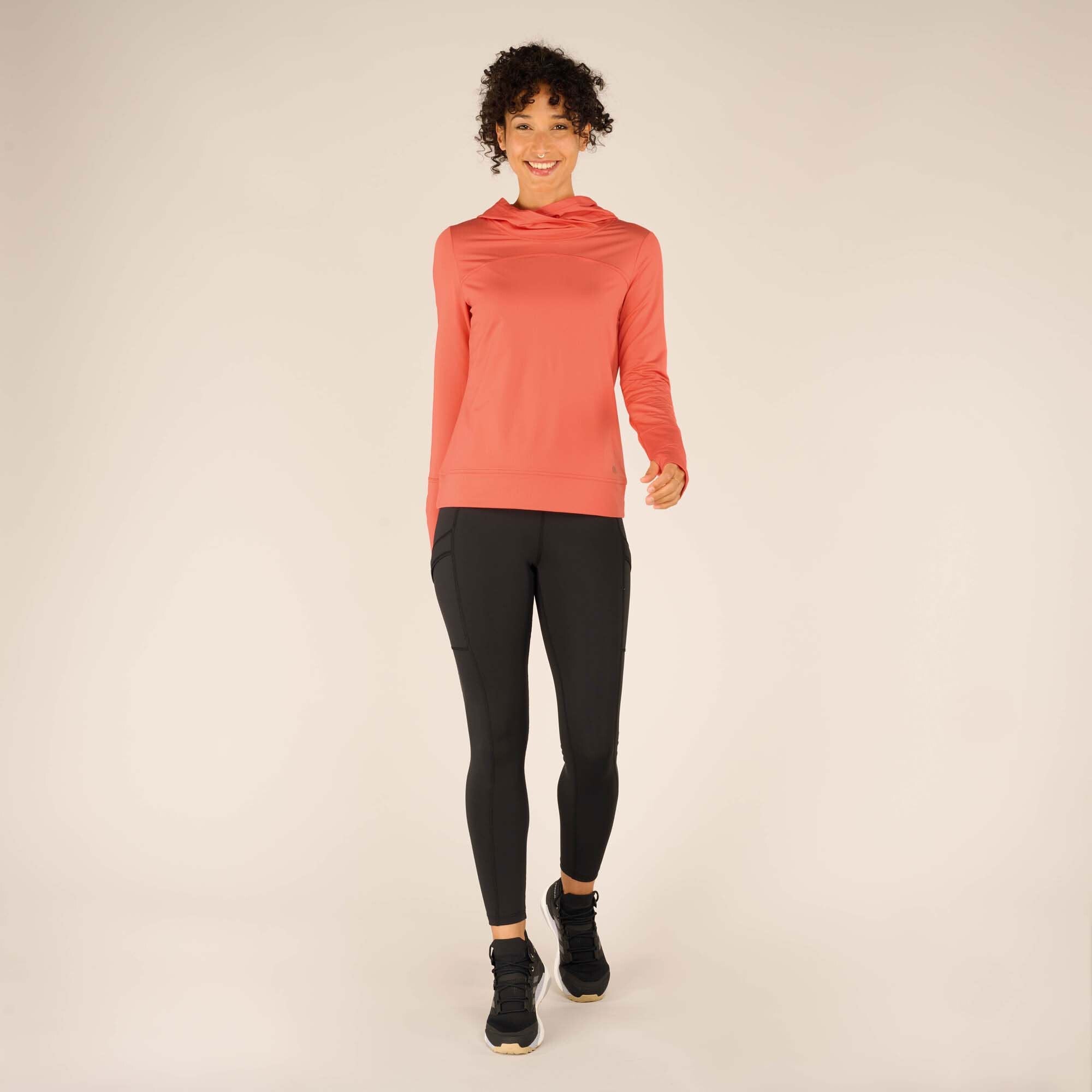 Full-body shot of the woman walking toward the camera wearing the Sherpa Adventure Gear Aarti Hoodie in Pink paired with black leggings and black sneakers. The hoodie extends slightly past her waist with a ribbed hem, and the sleeves feature thumbholes.