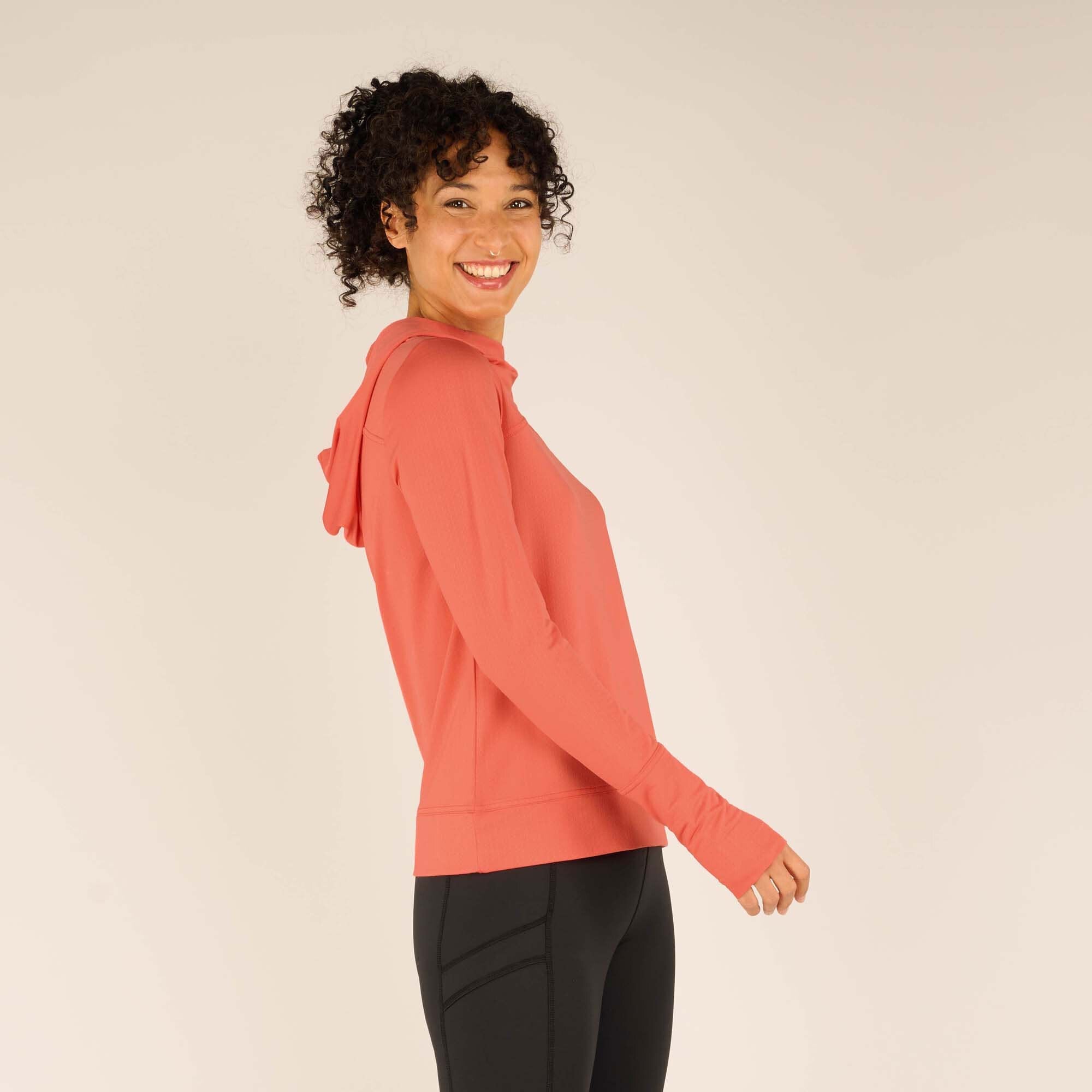 Side profile of the woman wearing the Sherpa Adventure Gear Aarti Hoodie in Pink, smiling at the camera. The fit is slightly loose, and the fabric drapes comfortably. The hood is down, resting on her back.