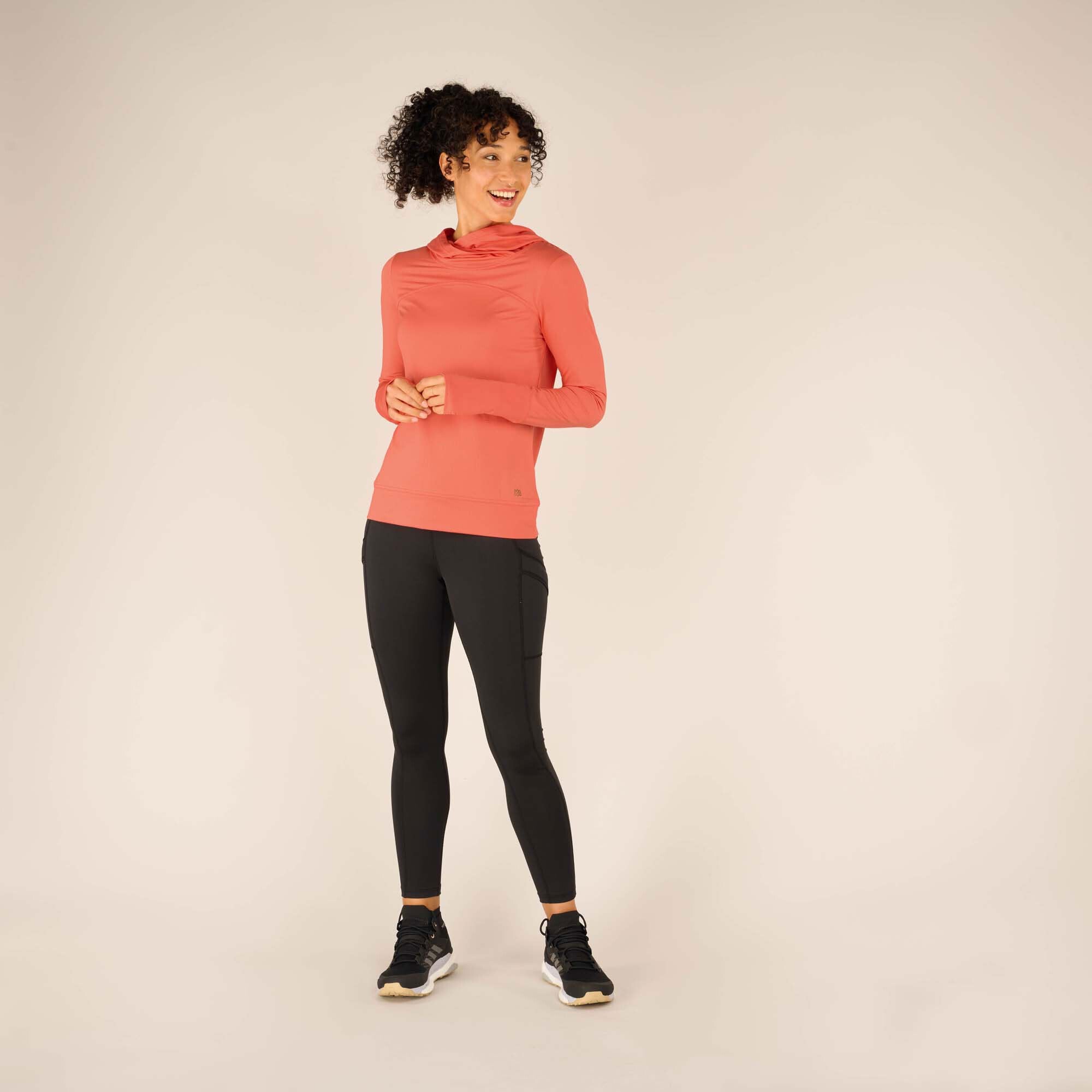 The woman stands in a relaxed pose with hands clasped together in front of her, wearing the Sherpa Adventure Gear Aarti Hoodie in Pink. The front seam detail is visible, adding subtle structure to the design.