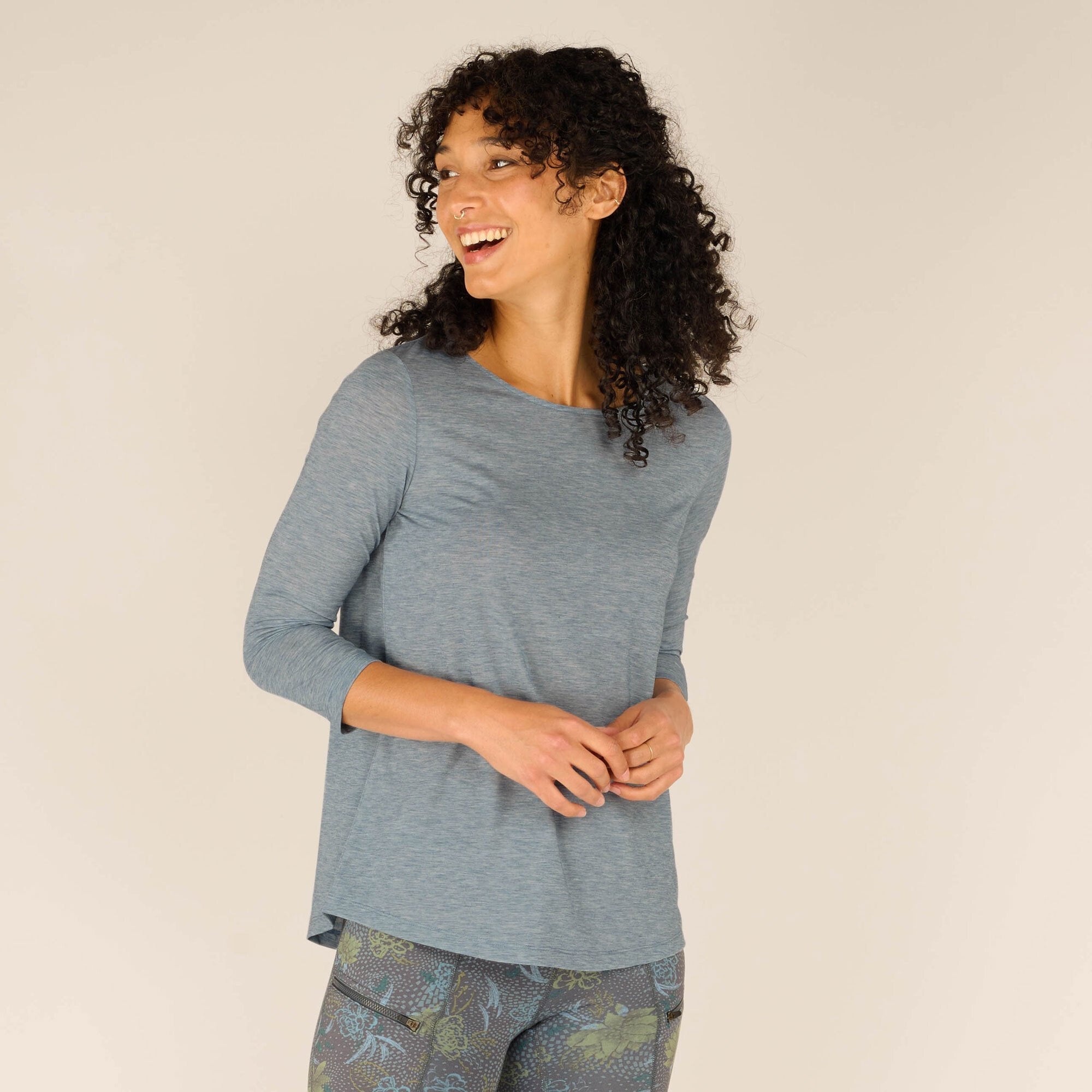 A smiling woman with curly hair wears a Sherpa Adventure Gear Asha 3/4 Sleeve Top in Blue paired with shorts featuring a nature-inspired floral pattern in blue and green hues. The photo is taken in a studio with a beige background, and the woman is slightly angled toward the camera while laughing.