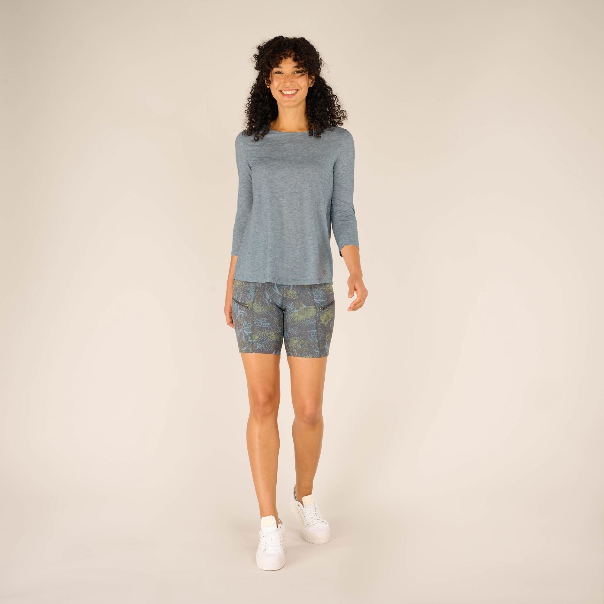 The same model is seen walking confidently toward the camera, showcasing the flowy fit of the Sherpa Adventure Gear Asha 3/4 Sleeve Top in Blue and the intricate details of the floral-patterned shorts. She pairs the outfit with white trainers, standing against a neutral beige background.
