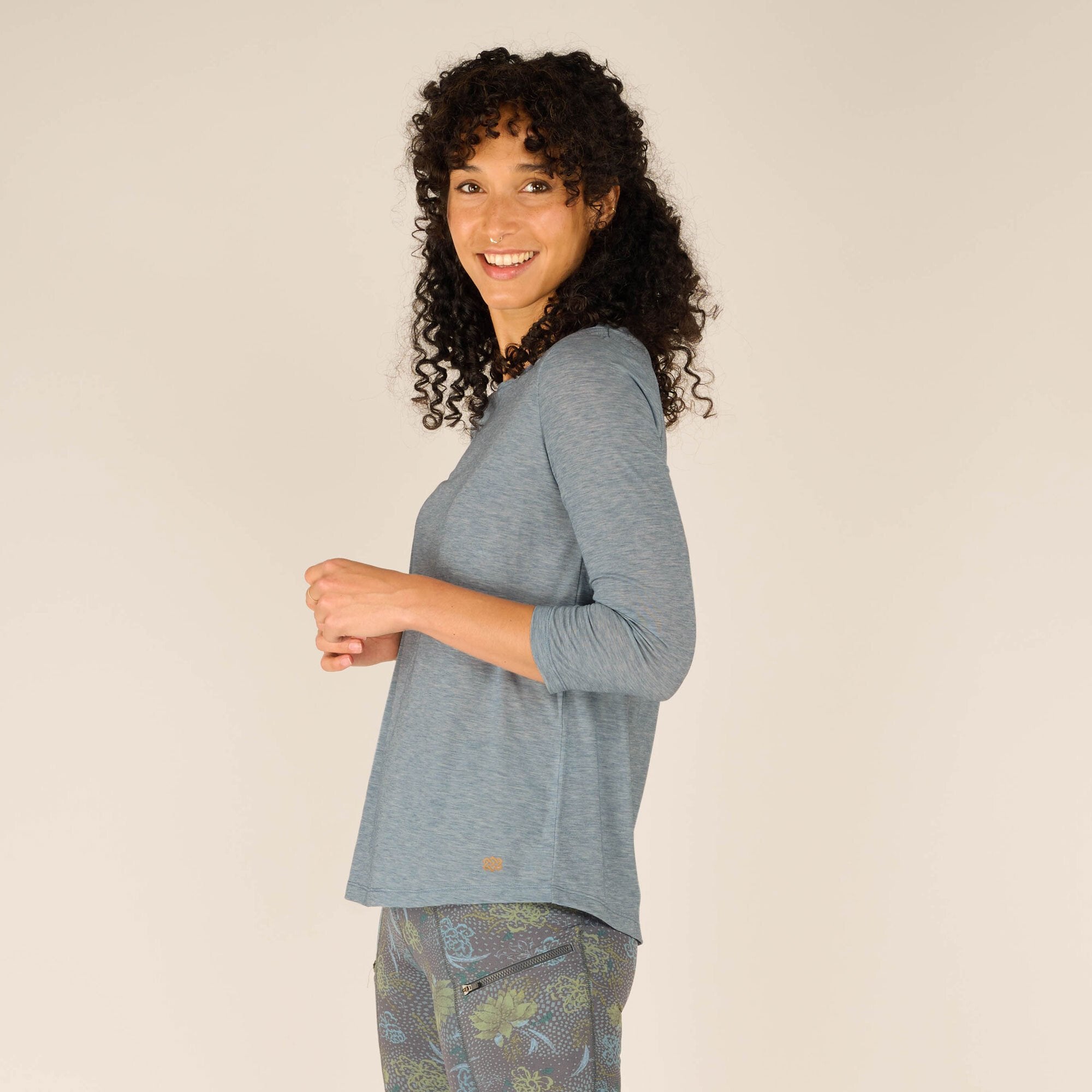 The model is in a side profile pose, turning slightly toward the camera. The loose and relaxed fit of the Sherpa Adventure Gear Asha 3/4 Sleeve Top in Blue is evident, with the floral-patterned shorts peeking through. Her cheerful expression highlights the casual yet stylish vibe of the outfit.