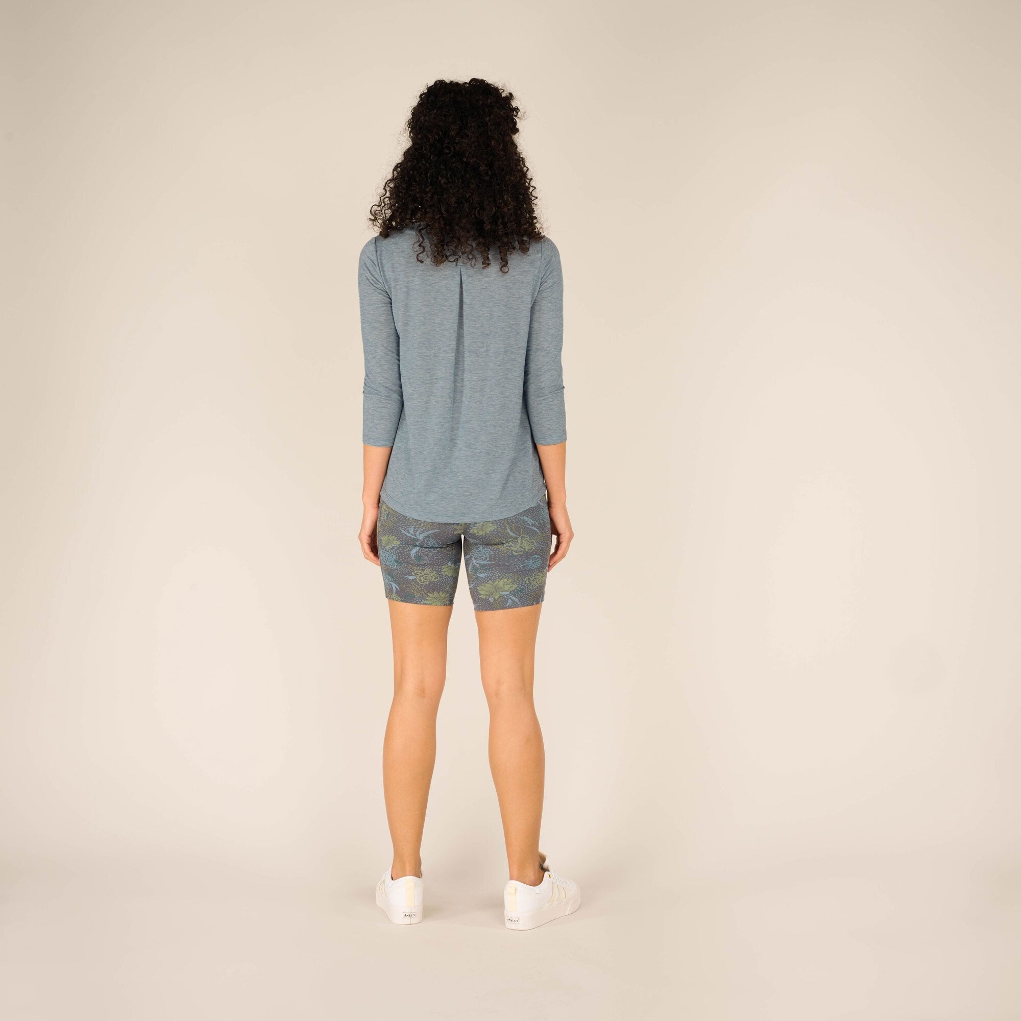 The model is captured from the back, displaying the draped back of the Sherpa Adventure Gear Asha 3/4 Sleeve Top in Blue and the rear design of the patterned shorts. The neutral beige studio setting highlights the vibrant colors and patterns of the clothing.