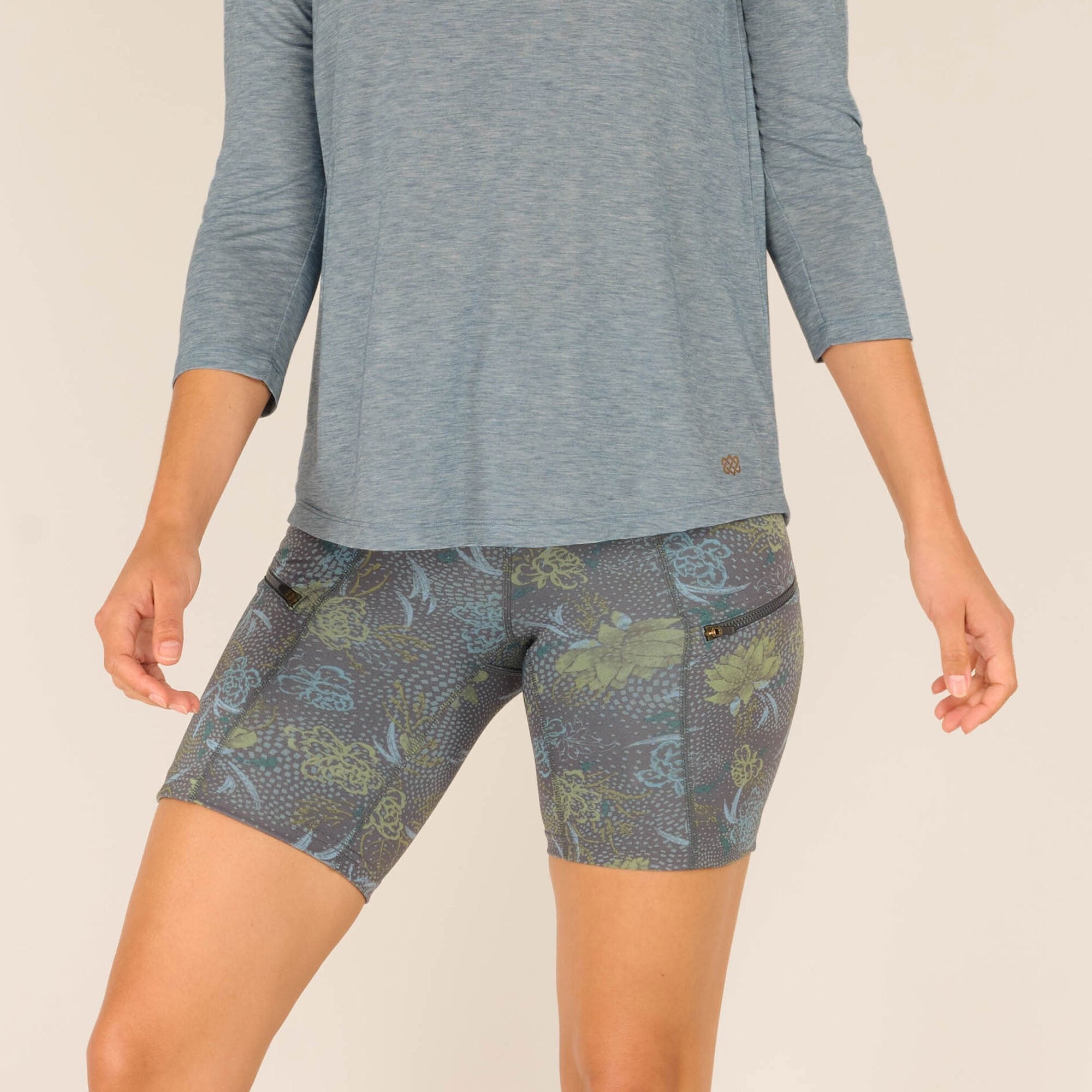 A close-up shot of the lower portion of the outfit, focusing on the intricate floral details of the shorts and the hemline of the Sherpa Adventure Gear Asha 3/4 Sleeve Top in Blue. The zippered pocket on the shorts adds a functional detail, while the floral design showcases a mix of blue and green botanical elements.