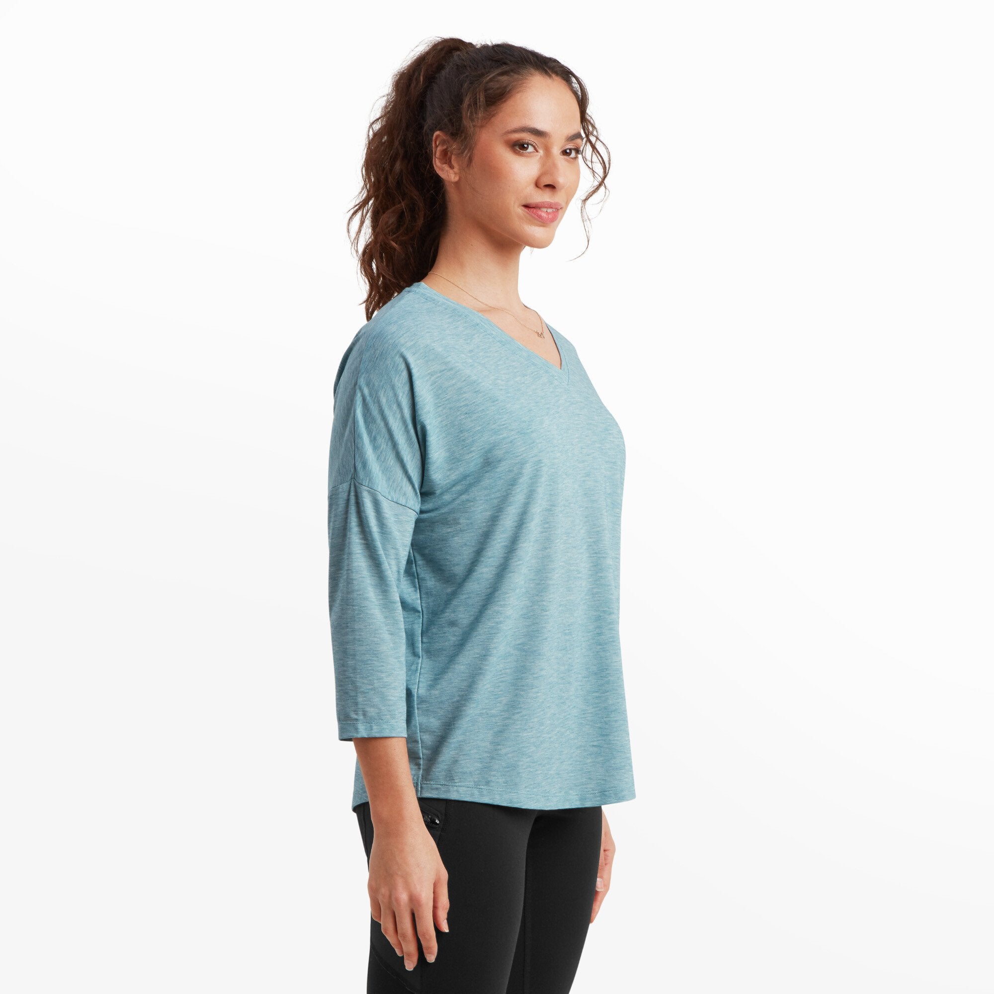 The model is turned slightly to the side, showcasing the Sherpa Adventure Gear Asha V-Neck 3/4 Sleeve Top in Blue's drape and relaxed silhouette. The V-neck and dropped shoulders are visible, and her left arm is relaxed at her side. Her hair is styled in a high ponytail, and she has a subtle smile on her face.