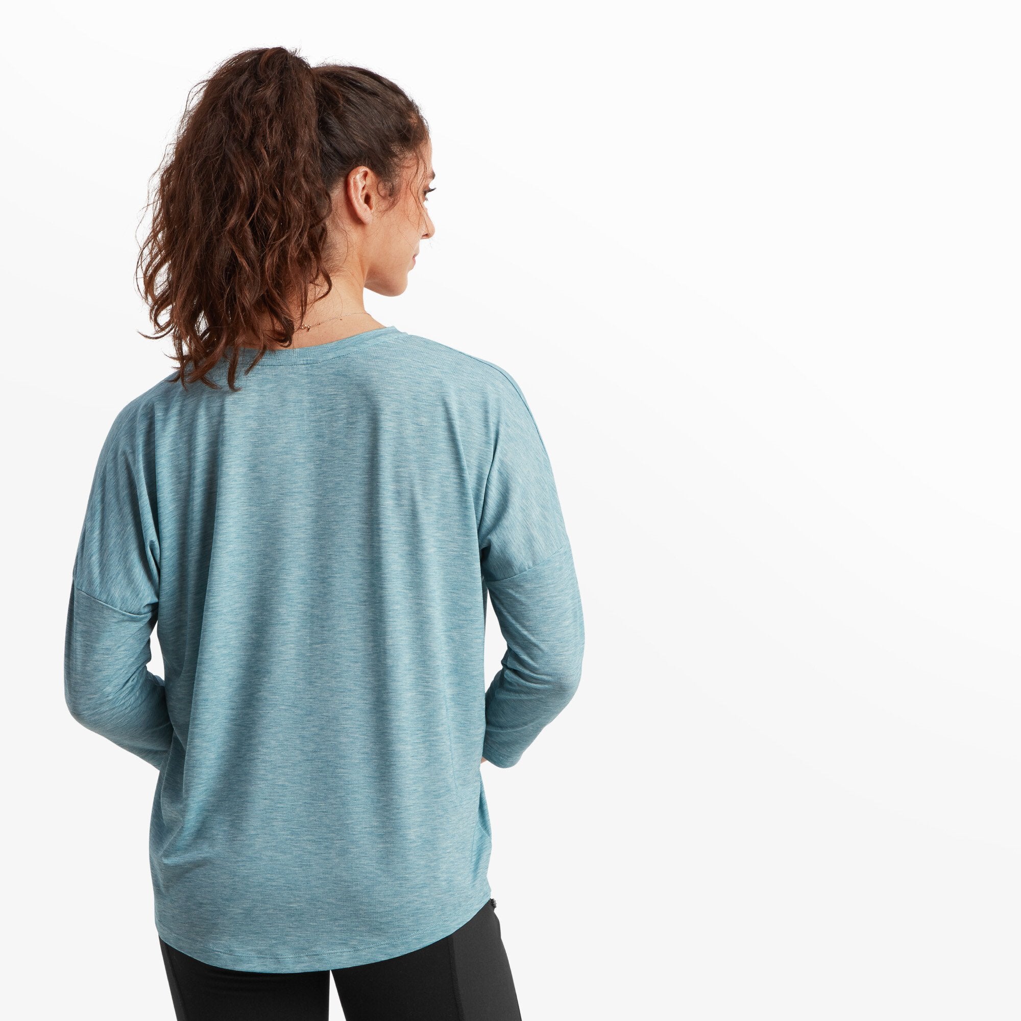 The model faces away from the camera, revealing the back of the Sherpa Adventure Gear Asha V-Neck 3/4 Sleeve Top in Blue. The fabric flows loosely over her frame, with a slightly rounded hemline. The heathered texture of the material is visible, and her wavy ponytail cascades down her back.