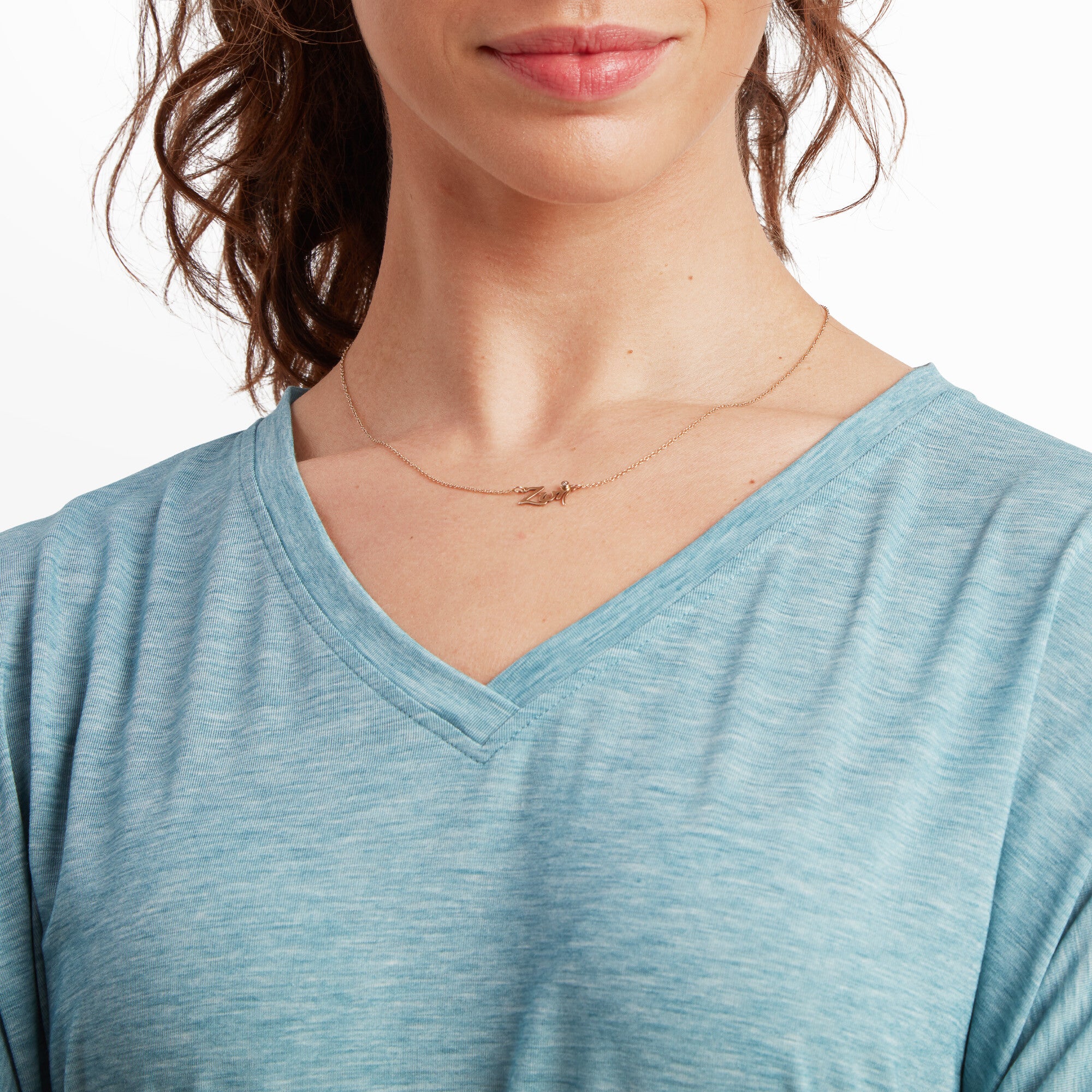 A zoomed-in image highlighting the V-neck cut of the Sherpa Adventure Gear Asha V-Neck 3/4 Sleeve Top in Blue, with visible stitching around the collar. The model is wearing a delicate gold necklace with a small pendant. The soft fabric texture is evident, and a portion of her lips and jawline are also visible in the frame.