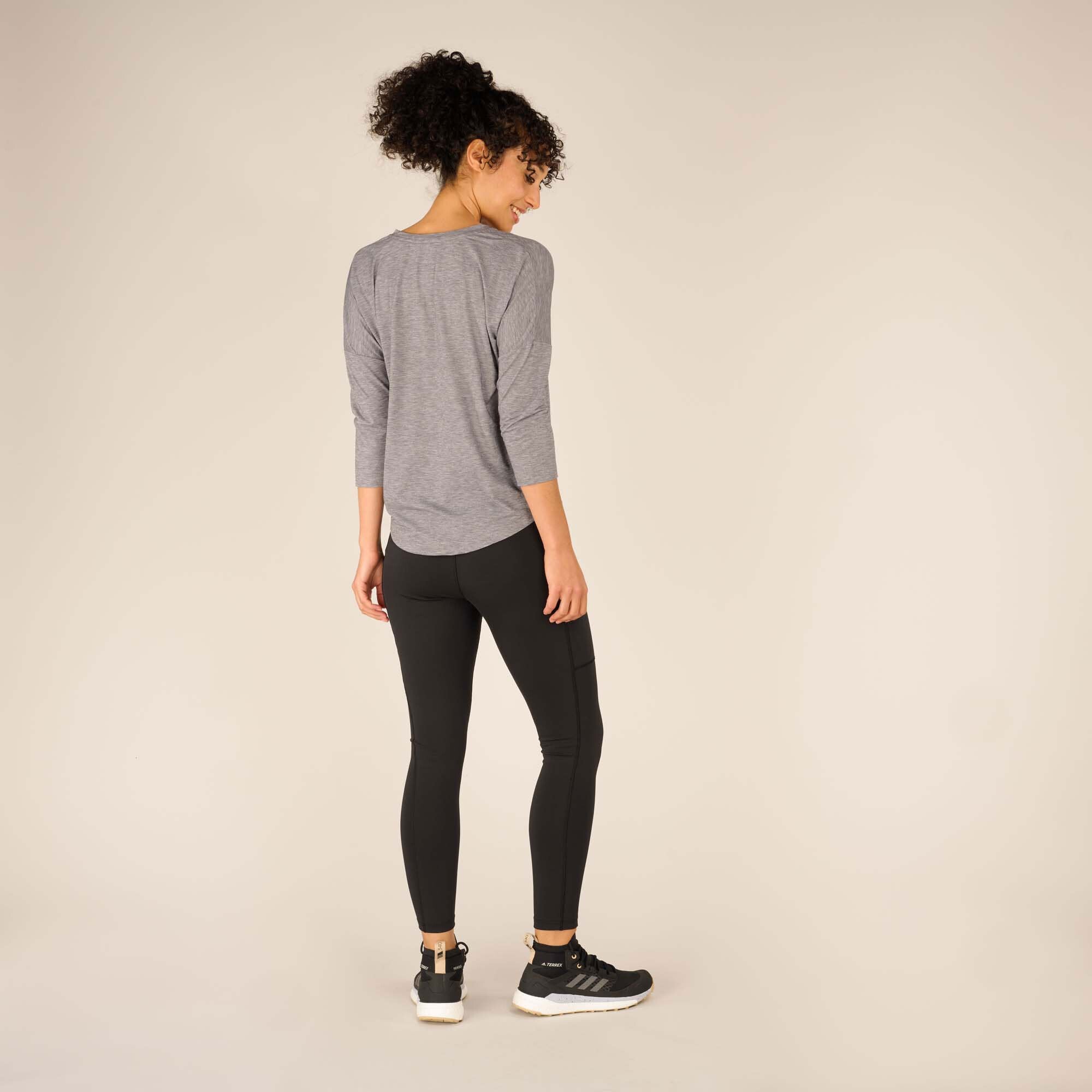 The woman is seen from behind, showcasing the flowy hemline of the Sherpa Adventure Gear Asha V-Neck 3/4 Sleeve Top in Grey. The subtle pleating in the back adds texture and dimension to the garment. Her black leggings and trainers enhance the outfit's activewear appeal, while her posture conveys a relaxed and confident demeanor.