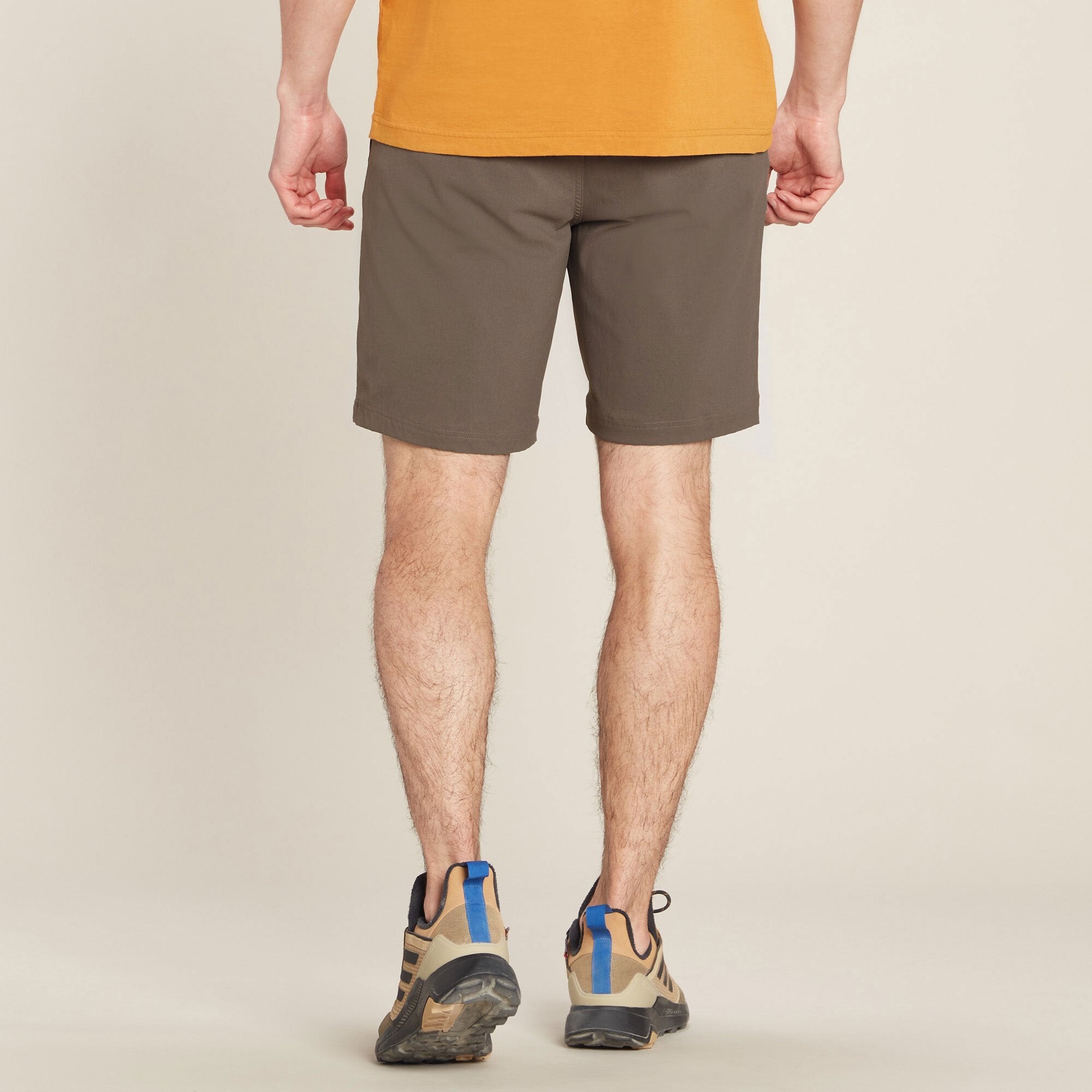The same male model is shown from the back, showcasing the rear fit of the Sherpa Adventure Gear Bara Shorts in Green. The waistband sits comfortably on his hips, and the shorts have a clean, structured look with no visible back pockets. The model's legs are slightly apart, and his hands rest naturally. The background remains a neutral beige.