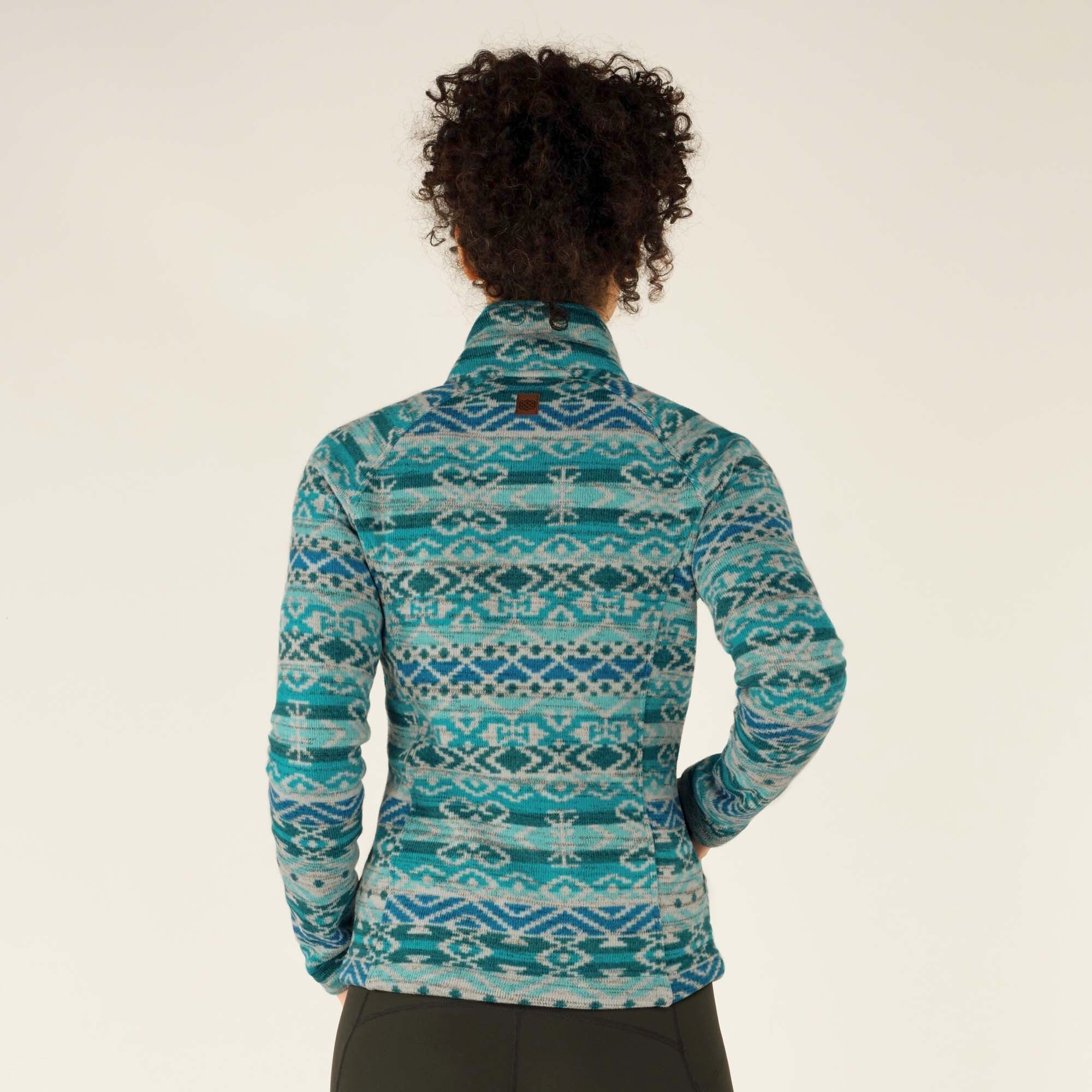 A back view of the model wearing the Sherpa Adventure Gear Bhutan Full Zip Jacket in Green. The symmetrical geometric pattern in shades of teal and blue covers the entire back. The high collar and tailored fit create a structured yet cozy look. A small brown leather brand logo patch is visible at the center of the upper back.