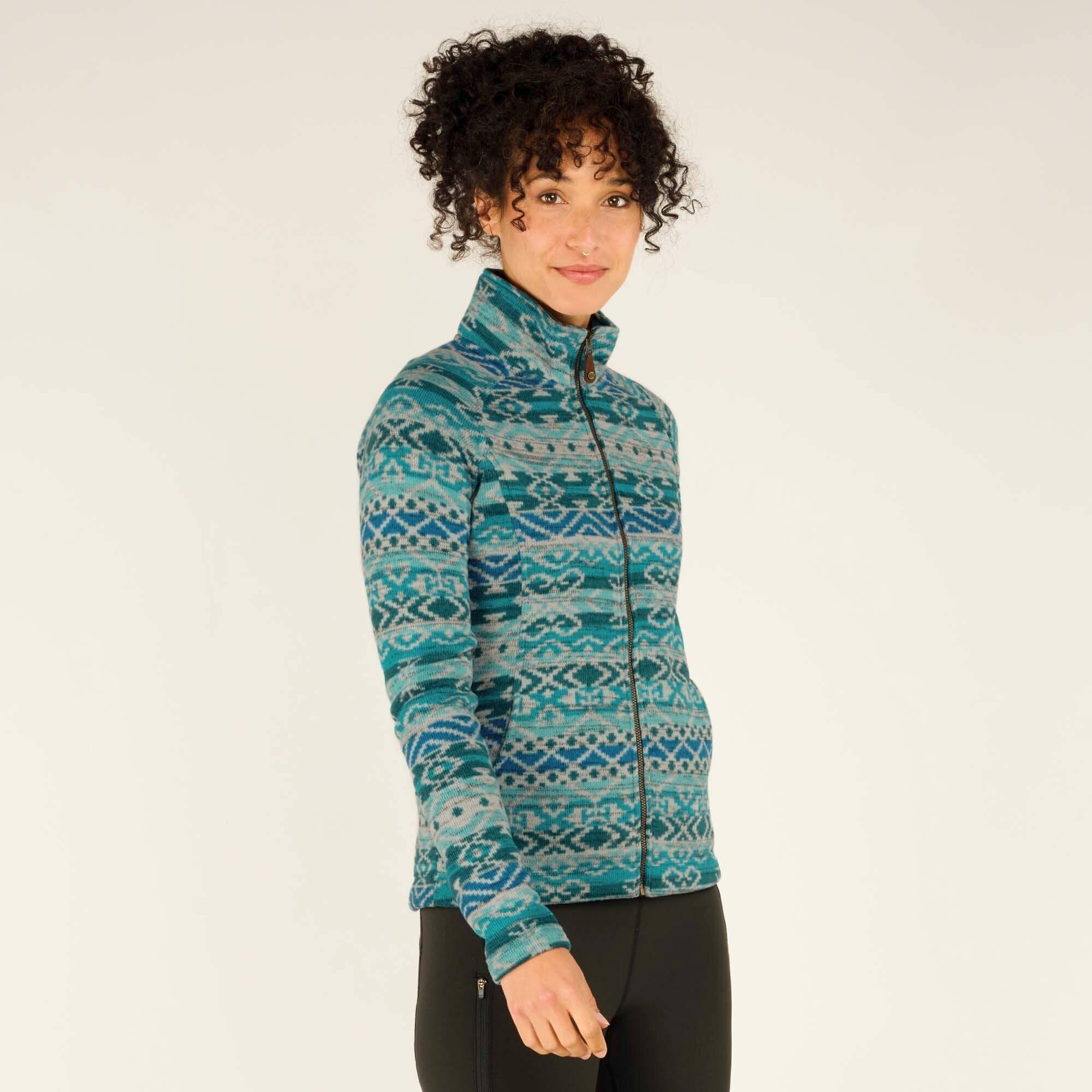 A front-facing shot of the model standing naturally while wearing the Sherpa Adventure Gear Bhutan Full Zip Jacket in Green. The full-length zipper, high collar, and side pockets are visible, and the model gazes at the camera with a calm expression. The jacket’s pattern is intricate and symmetrical, blending traditional motifs with modern styling.