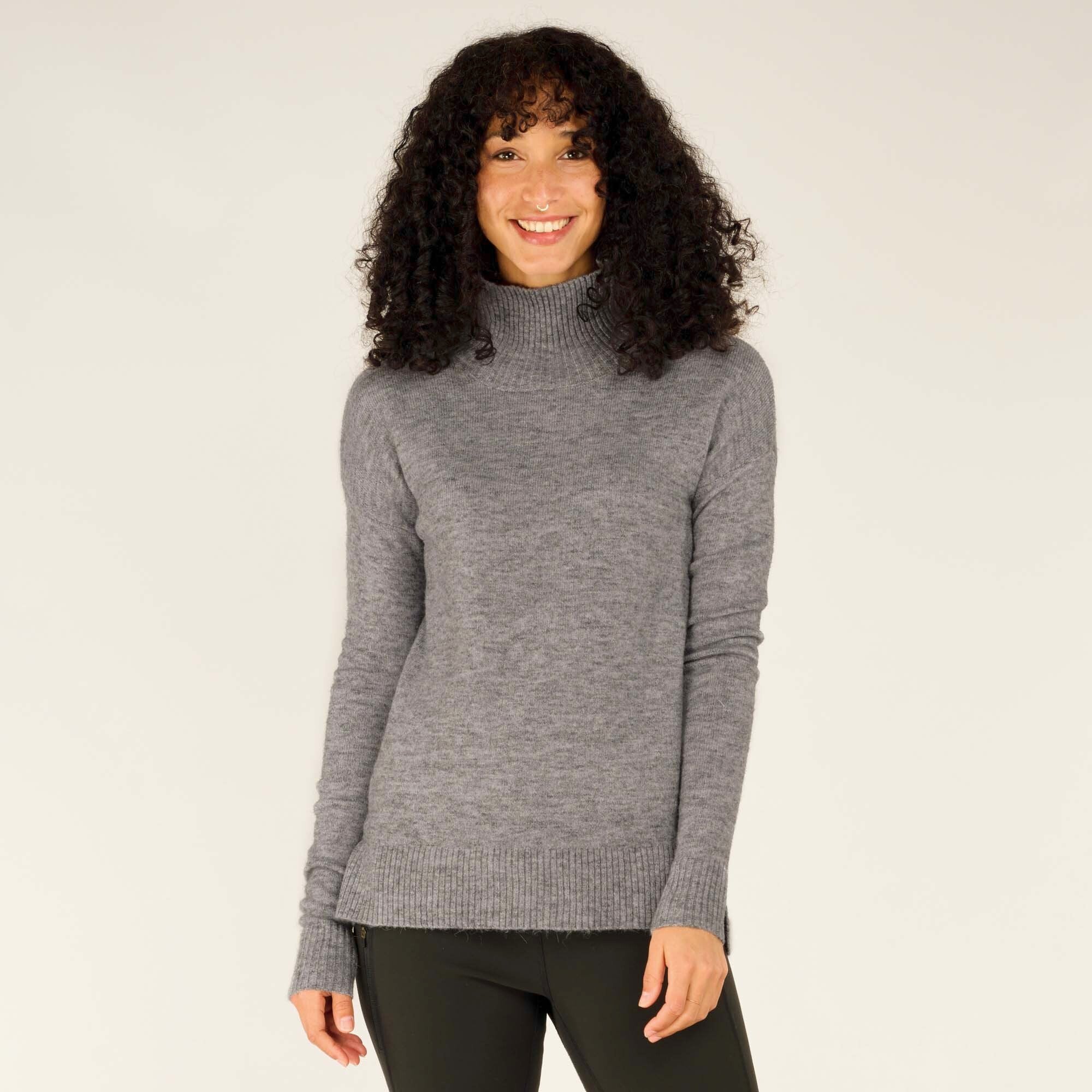 A model is wearing a Sherpa Adventure Gear Dali Mock Neck in Grey made of soft material. The sweater has a ribbed texture at the neckline, cuffs, and hem, offering a snug and stylish fit. The long sleeves extend slightly over the wrists, giving a cozy look. The sweater is paired with dark green slim-fit pants and the model is smiling brightly at the camera.