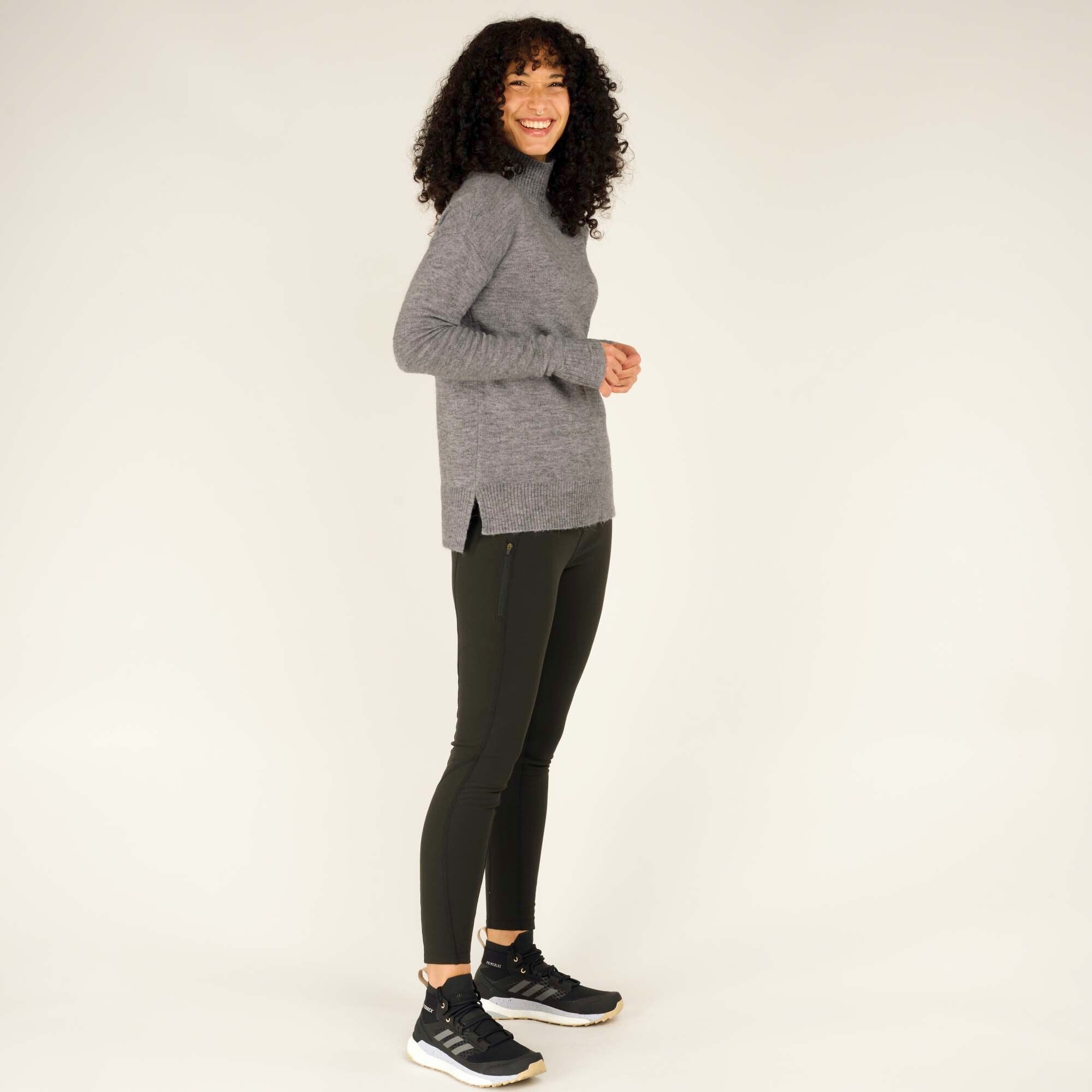 The model is showcasing the side profile of the Sherpa Adventure Gear Dali Mock Neck in Grey. The hem has a small slit on each side, adding a touch of detail and allowing for better movement. The texture and fine-knit quality of the sweater are visible. The model is paired with the same dark green pants and black athletic shoes.