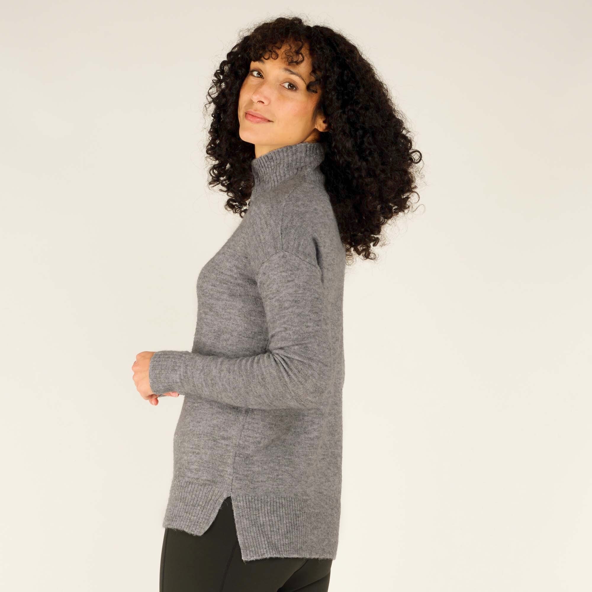 A close side view highlights the snug fit of the Sherpa Adventure Gear Dali Mock Neck in Grey along the shoulders and arms, as well as the soft flow of the fabric at the hemline. The high neckline is seen fitting closely to the neck, adding to the warmth and design. The model gazes softly at the camera, emphasising the sweater’s elegant simplicity.