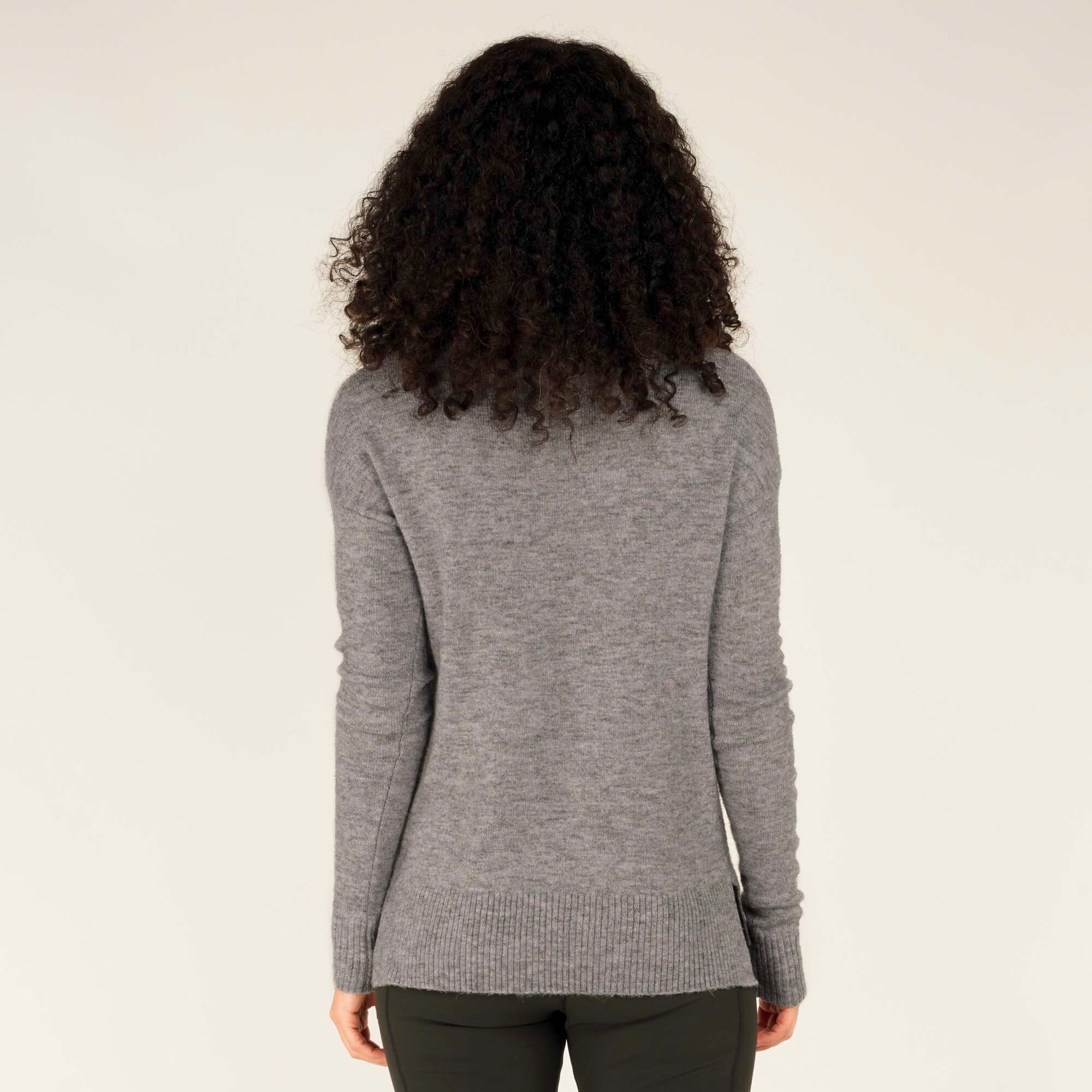 The back of the Sherpa Adventure Gear Dali Mock Neck in Grey is shown, featuring a plain design with a subtle ribbed pattern at the hem and cuffs. The model's hair flows naturally down her back, further emphasising the clean and classic design of the sweater.