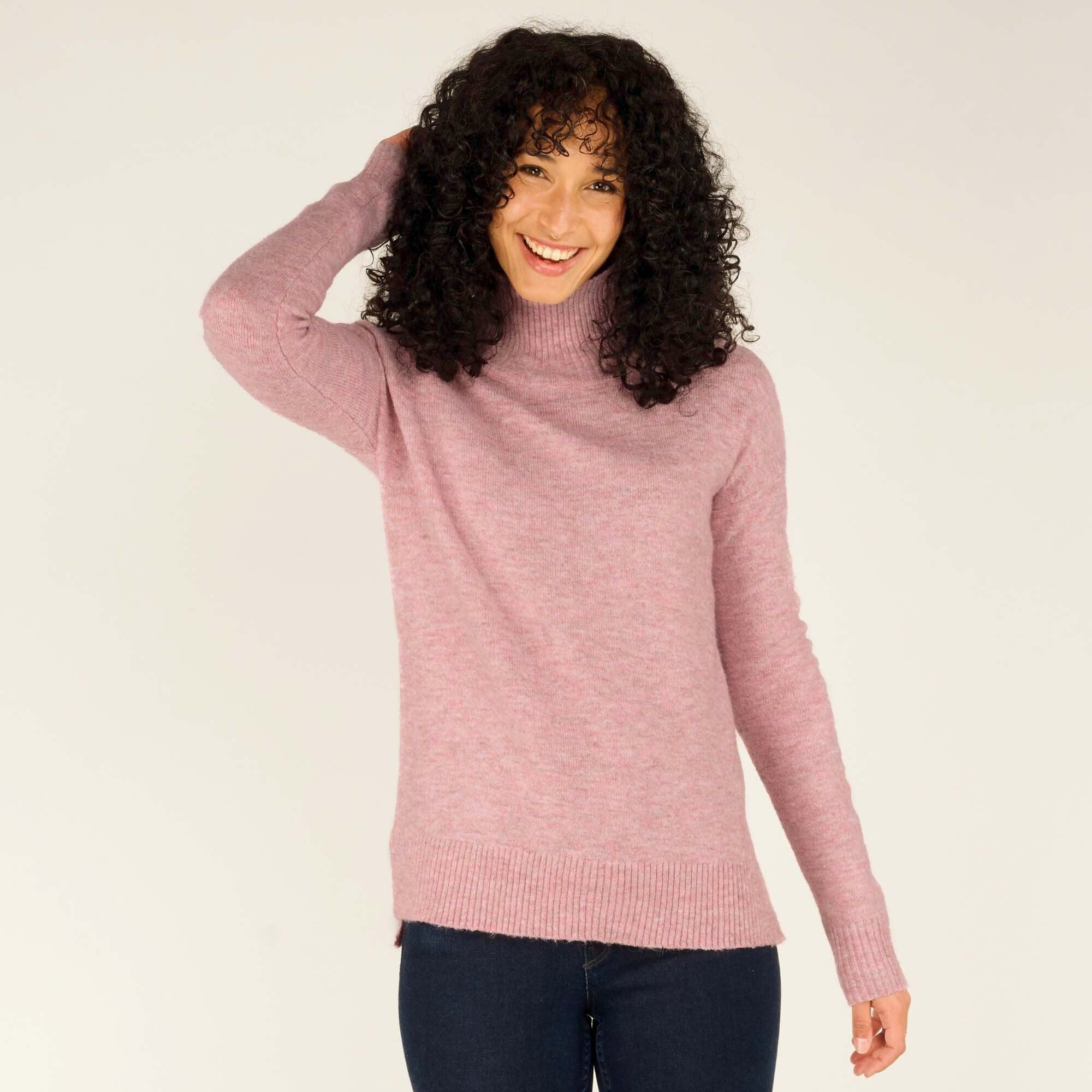 A woman smiling warmly, wearing a Sherpa Adventure Gear Dali Mock Neck in Pink with long sleeves. The sweater has a relaxed fit and features a subtle ribbed hemline at the bottom. The model's curly hair frames her face as she poses casually with one hand lifting her hair. She is paired with dark skinny jeans for a cozy yet stylish look.