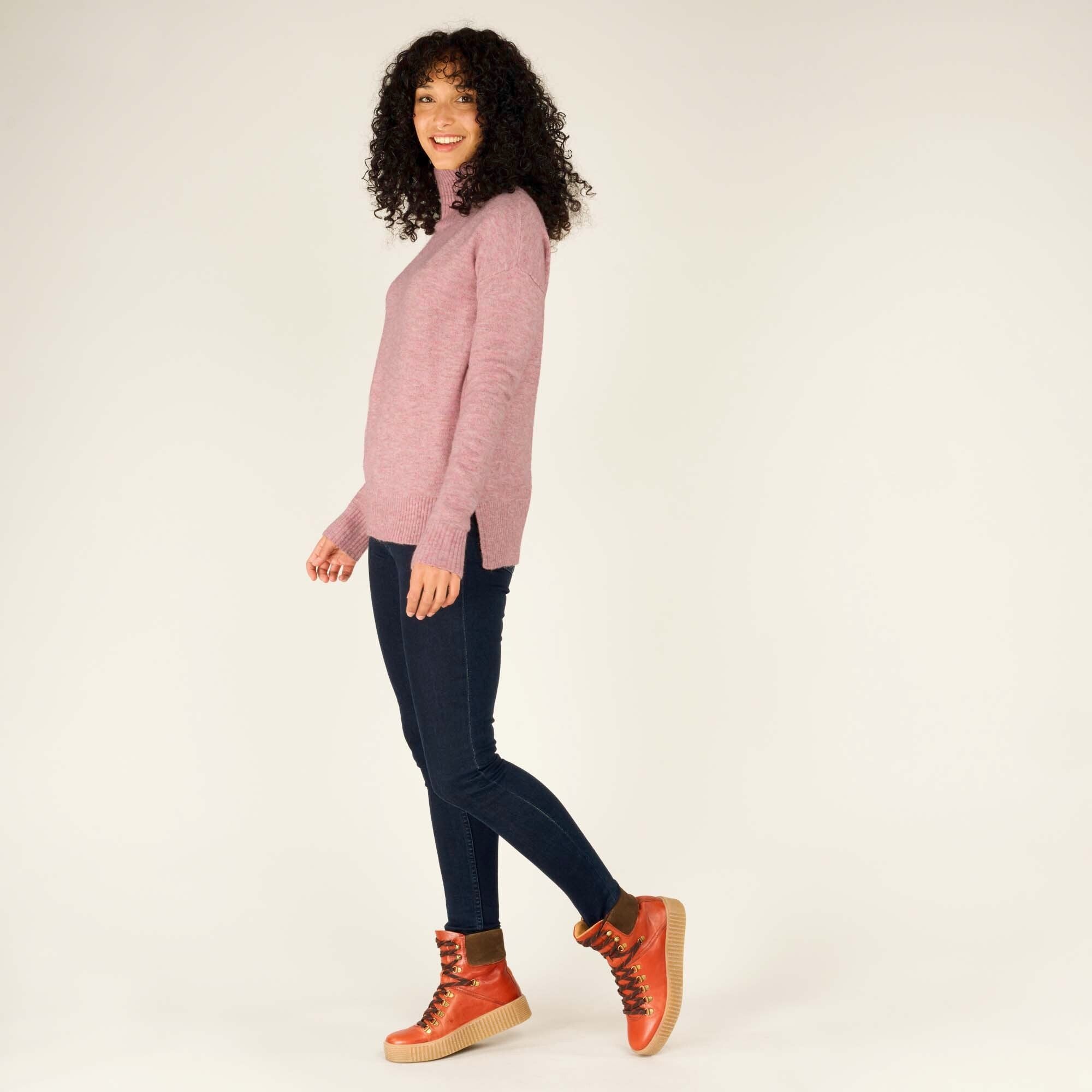 The same model takes a side step in profile view, showcasing the Sherpa Adventure Gear Dali Mock Neck in Pink's side slits and hemline that extends slightly longer in the back. The soft pink mock neck contrasts beautifully with her dark skinny jeans and orange hiking boots. Her bright smile complements the warm and casual aesthetic of the outfit.