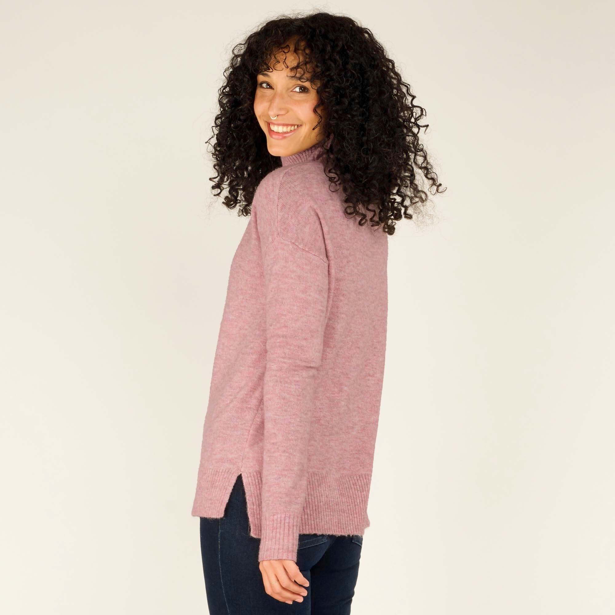 A three-quarter view of the woman looking over her shoulder, highlighting the soft drape and texture of the Sherpa Adventure Gear Dali Mock Neck in Pink's back. The sweater flows gently, maintaining its casual and relaxed fit, with her jeans and boots visible as part of the outfit.