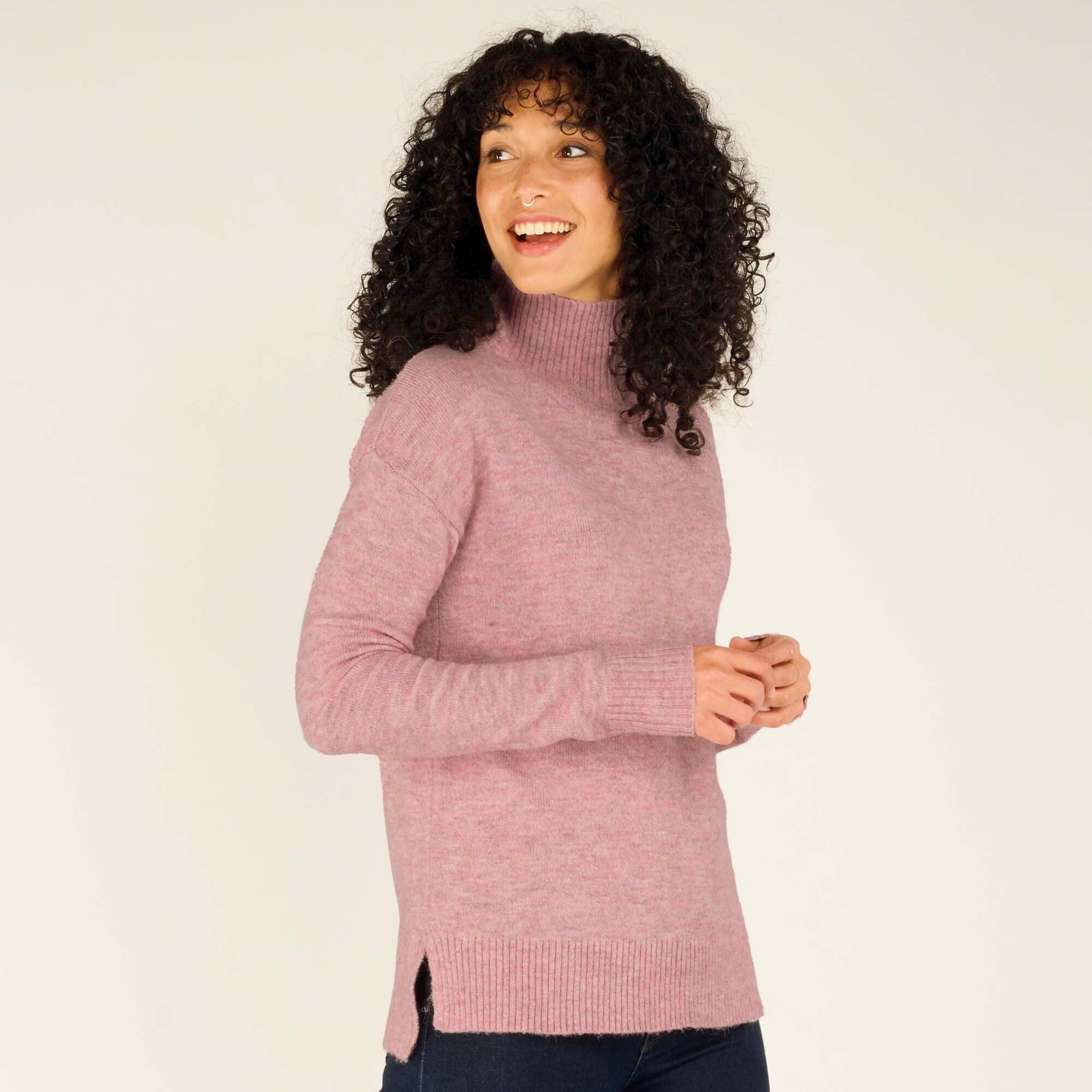 The woman faces the camera at a slight angle, smiling and clasping her hands together. The Sherpa Adventure Gear Dali Mock Neck in Pink's ribbed cuffs and neckline add structure to its cozy, knit design. The side slit detail can also be seen, paired perfectly with dark jeans and warm footwear.