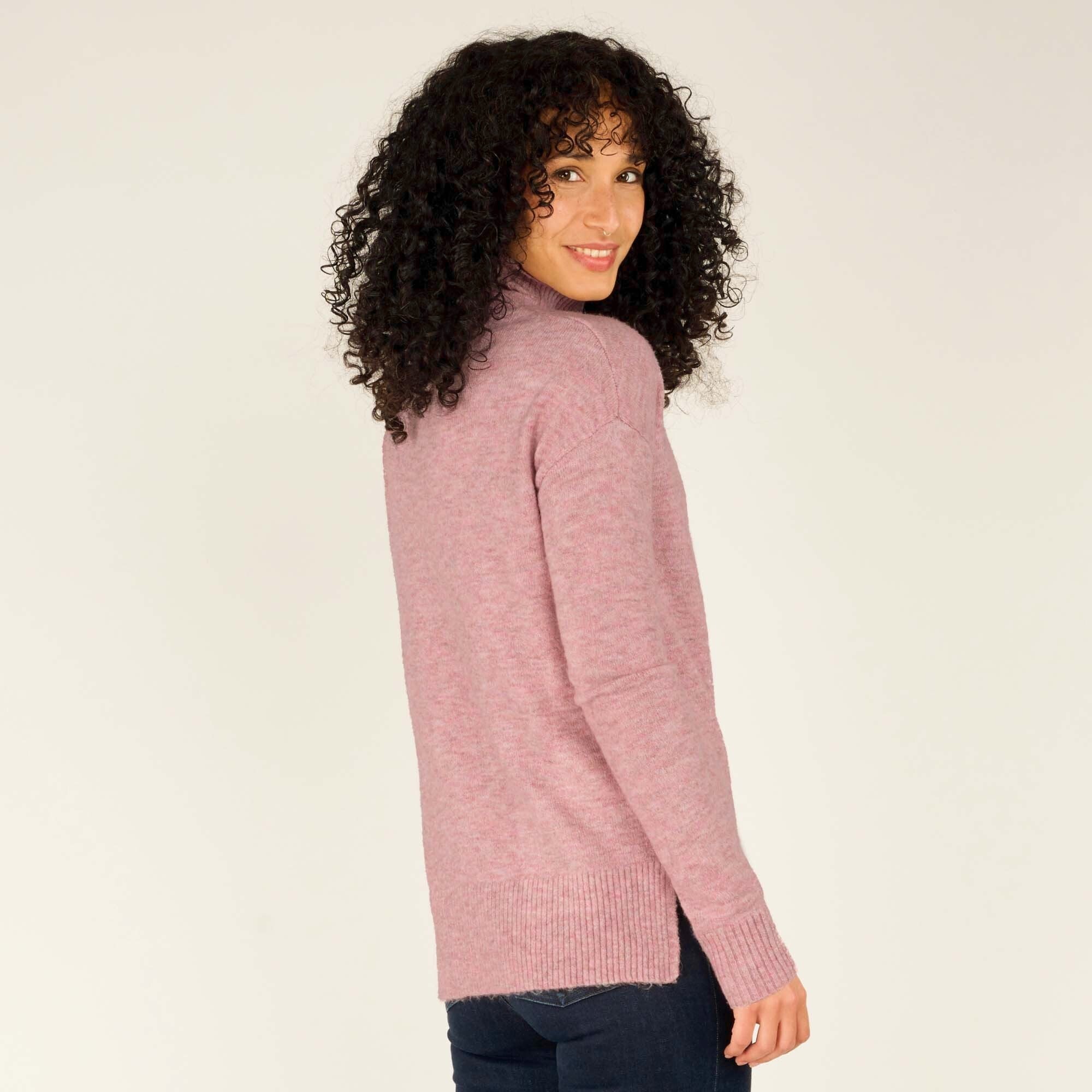 A back view of the model standing straight, her arms resting naturally by her sides. The Sherpa Adventure Gear Dali Mock Neck in Pink's simple design is shown from the back, emphasising the slightly longer hemline and subtle knit texture. The warm pink tone continues to stand out against her dark jeans and orange boots.