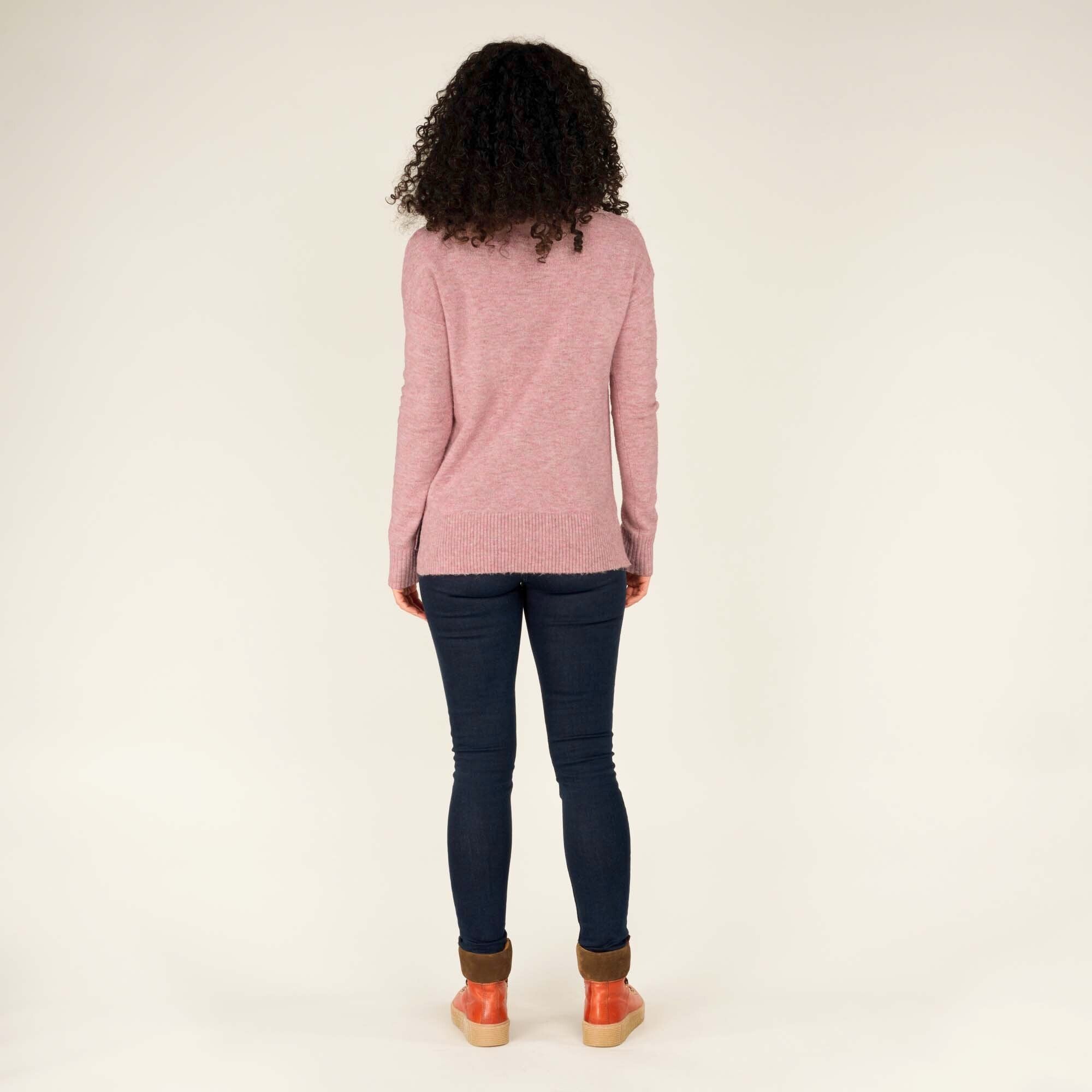 A full-length back profile of the model, allowing the entire outfit to be seen. The Sherpa Adventure Gear Dali Mock Neck in Pink, paired with dark skinny jeans and contrasting orange boots, creates a balanced look, ideal for casual and comfortable styling. The texture and lightweight feel of the sweater are emphasised through the model’s relaxed posture.