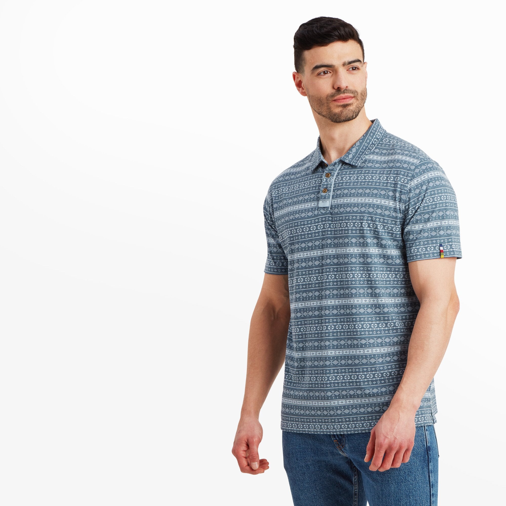 The same man is pictured in the Sherpa Adventure Gear Dhani Polo Shirt in Blue, standing at a slight angle with his head turned to the side, showcasing the shirt's fit across the shoulders and chest. He maintains a relaxed expression and stance, with his arms at his sides.