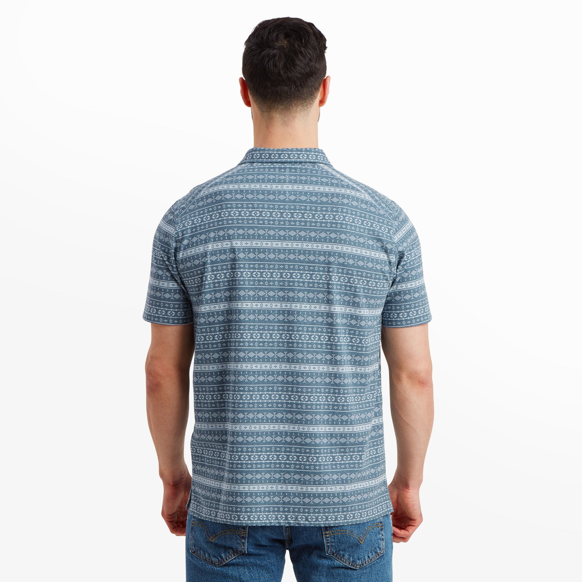 A back view of the man wearing the Sherpa Adventure Gear Dhani Polo Shirt in Blue, displaying the full geometric pattern across the back. The shirt fits comfortably, with clean, even lines. The man is standing upright, and the shirt is paired with blue jeans.