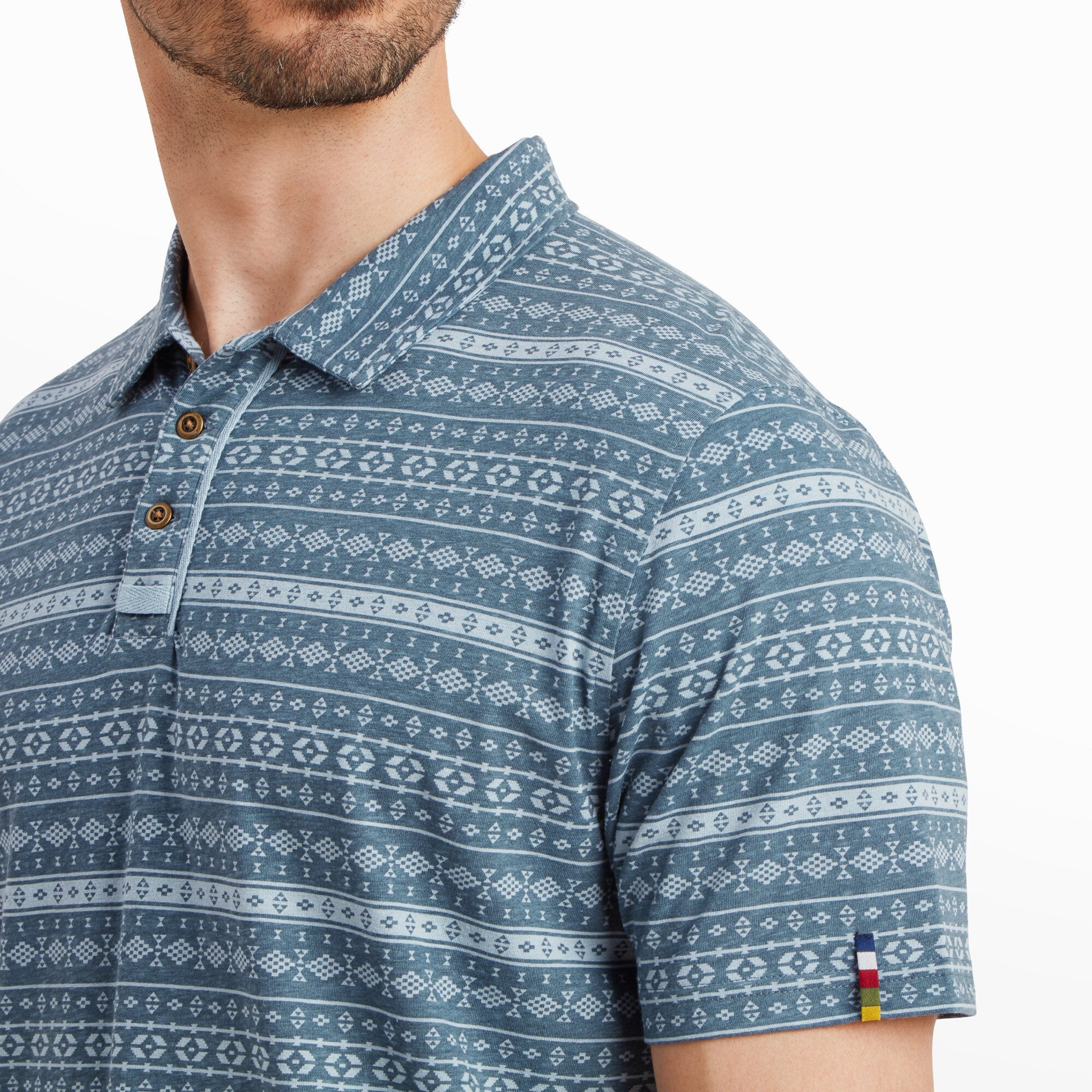 A detailed view of the Sherpa Adventure Gear Dhani Polo Shirt in Blue’s collar and upper chest area, highlighting the intricate geometric pattern and the wooden buttons on the placket. A small, colorful flag tag is visible on the sleeve hem, adding a unique design element. The stitching and fabric texture are clearly visible in this close-up.