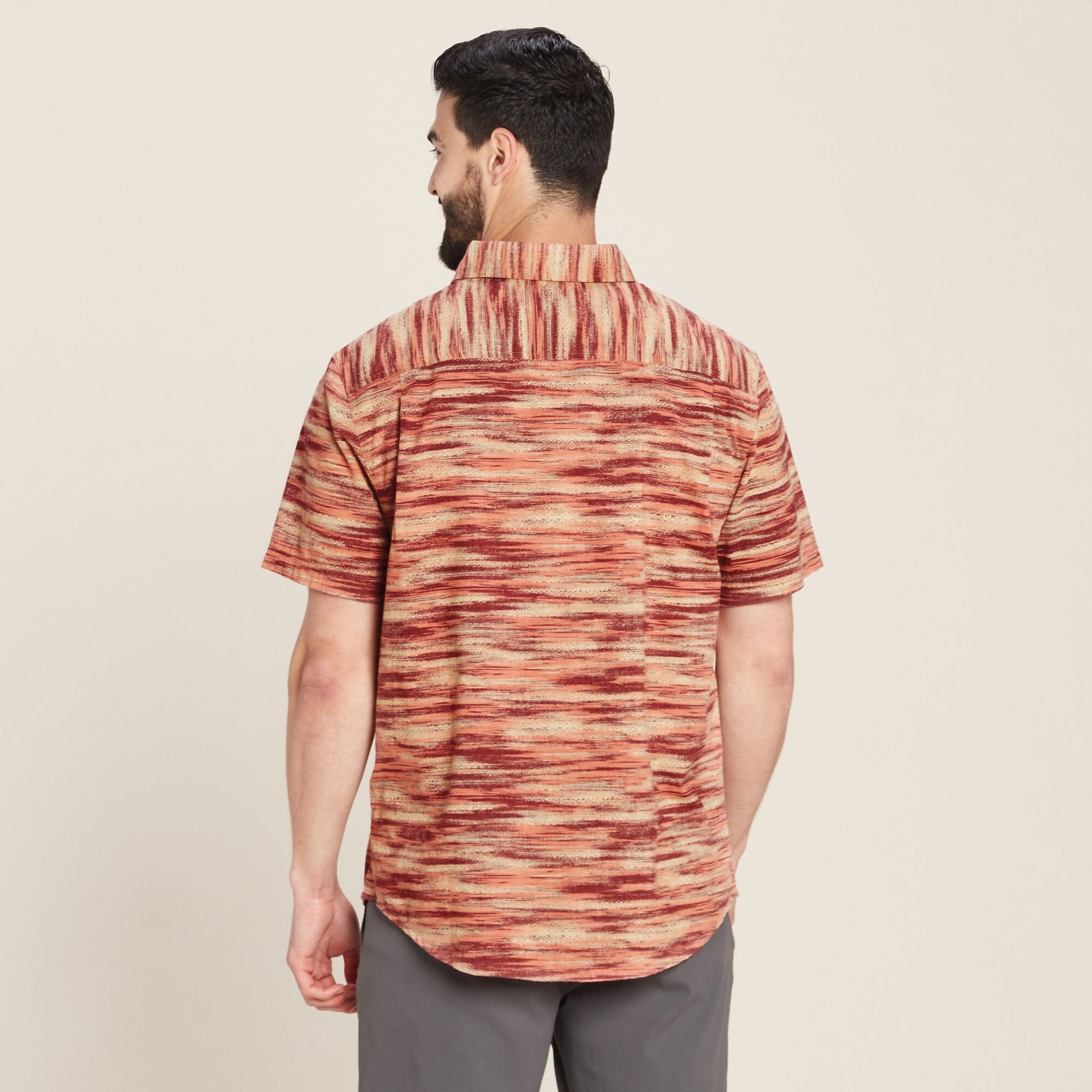 A rear perspective of the same Sherpa Adventure Gear Khamsu Short Sleeve Shirt in Beige, highlighting the continuous flow of the bold beige and red striped pattern across the back panel. The shirt maintains a relaxed fit, with its hem curving slightly at the bottom. The model's posture provides a clear view of the shirt's structure and design from the back.