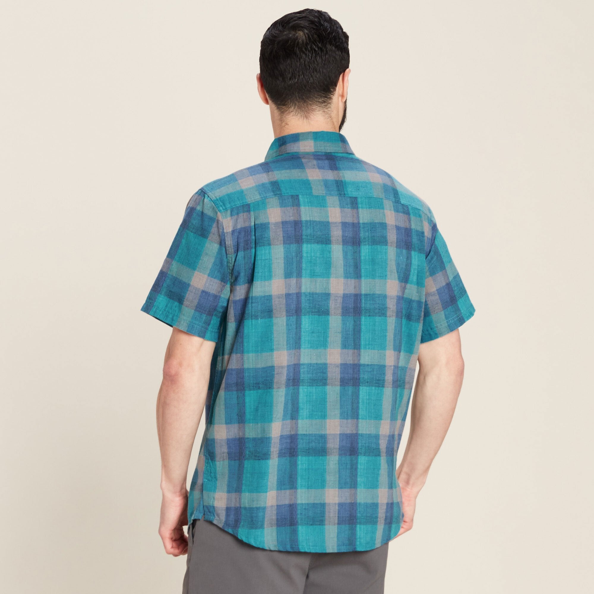 A rear view of the Sherpa Adventure Gear Khamsu Short Sleeve Shirt in Blue, showcasing its clean lines and slightly curved hem. The man pairs it with grey pants, and the light background highlights the shirt's vibrant colors.