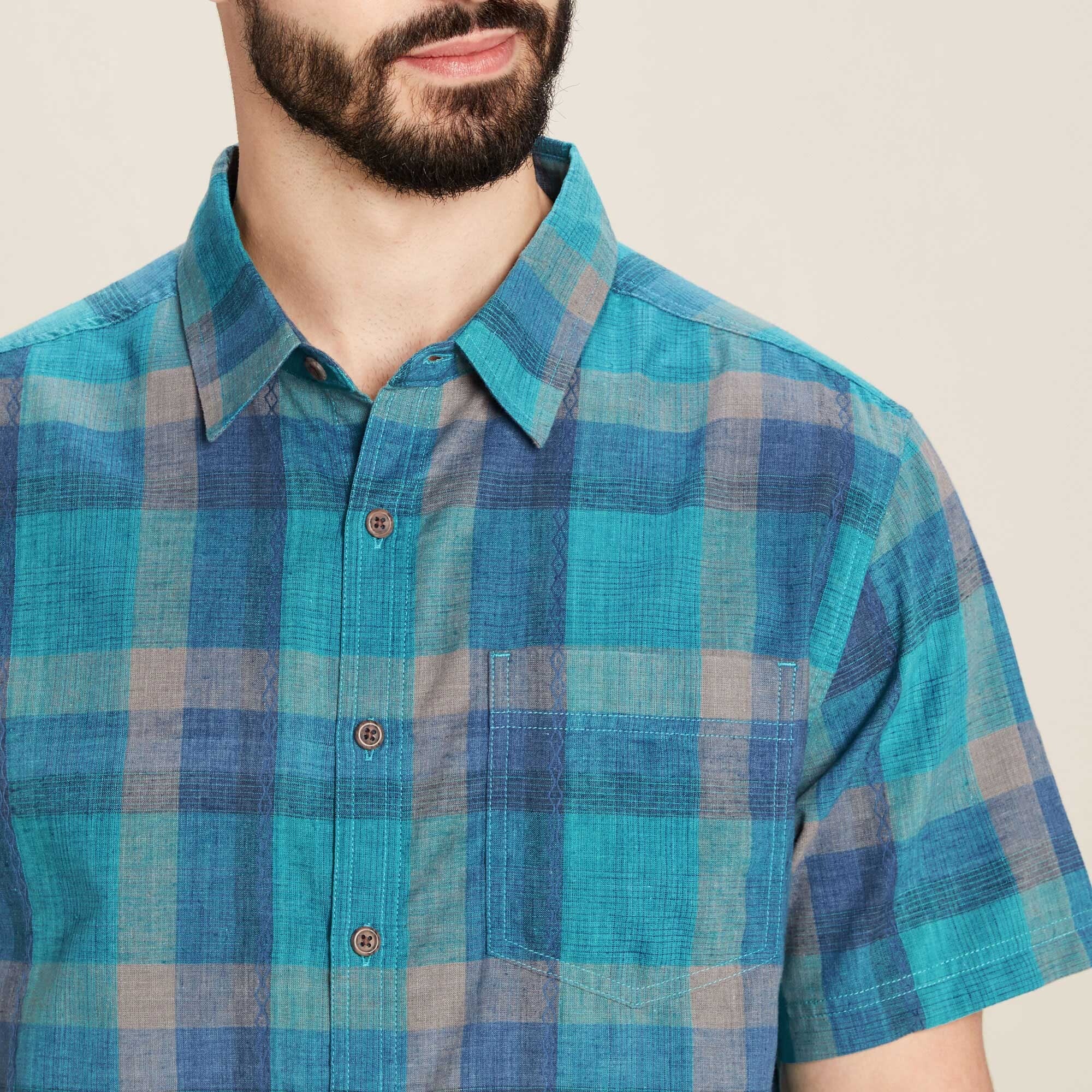 A close-up of the Sherpa Adventure Gear Khamsu Short Sleeve Shirt in Blue, focusing on the chest area. The shirt's texture and fine stitching details, including the buttons and single chest pocket, are clearly visible. The man stands against a neutral background, enhancing the shirt's vibrant checkered pattern.