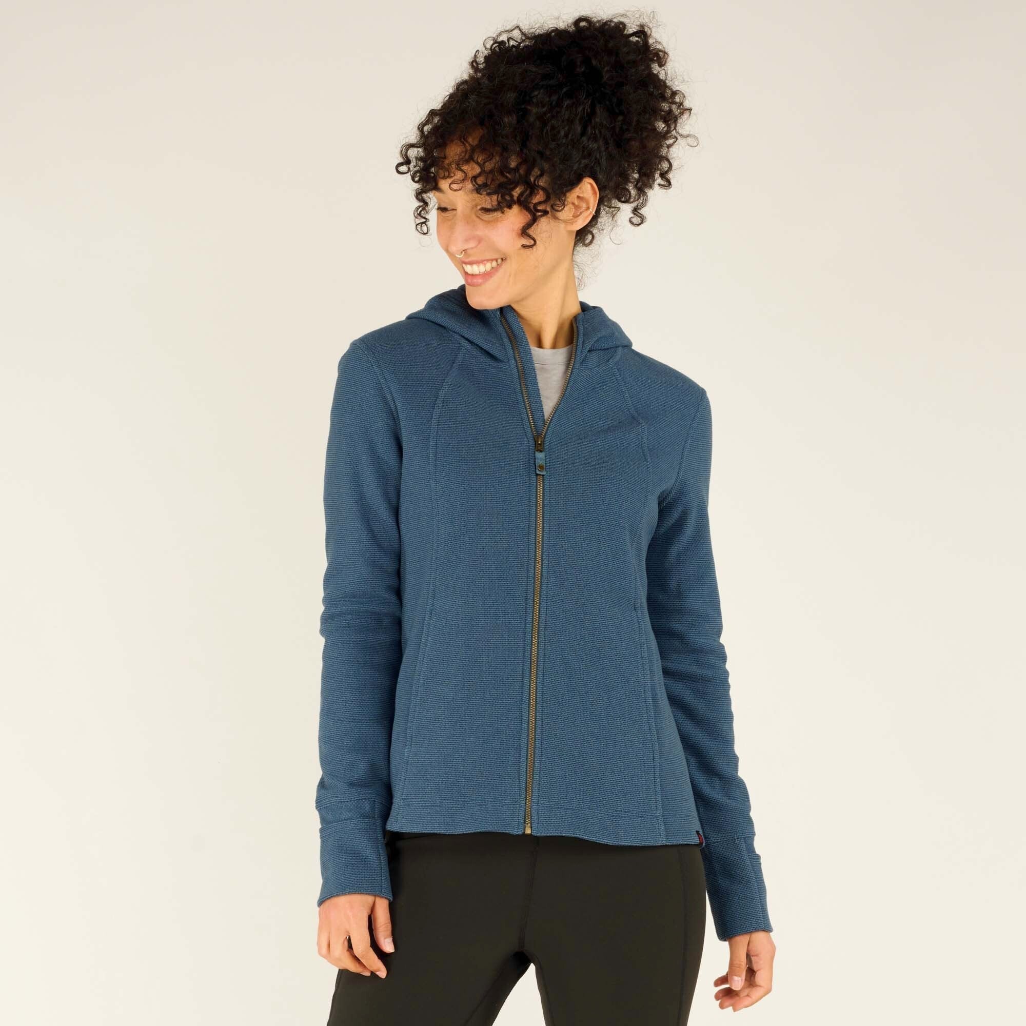 The same woman is seen standing upright, facing slightly to the side, smiling. The Sherpa Adventure Gear Kula Full Zip Hoodie in Blue is fully zipped up, and the hood rests against her back. Her black leggings and relaxed stance highlight the casual, comfortable nature of the outfit.