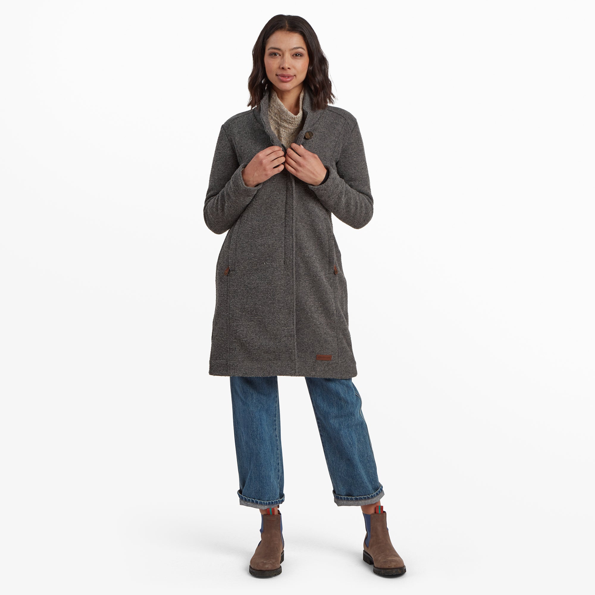 A woman with medium-length dark hair and a warm smile models a Sherpa Adventure Gear Lhamu Jacket in Grey. The coat features a structured collar with a single button closure near the neck, and vertical seams running down the front. She pairs the coat with a beige sweater, blue straight-leg jeans, and brown ankle boots with blue elastic side panels.