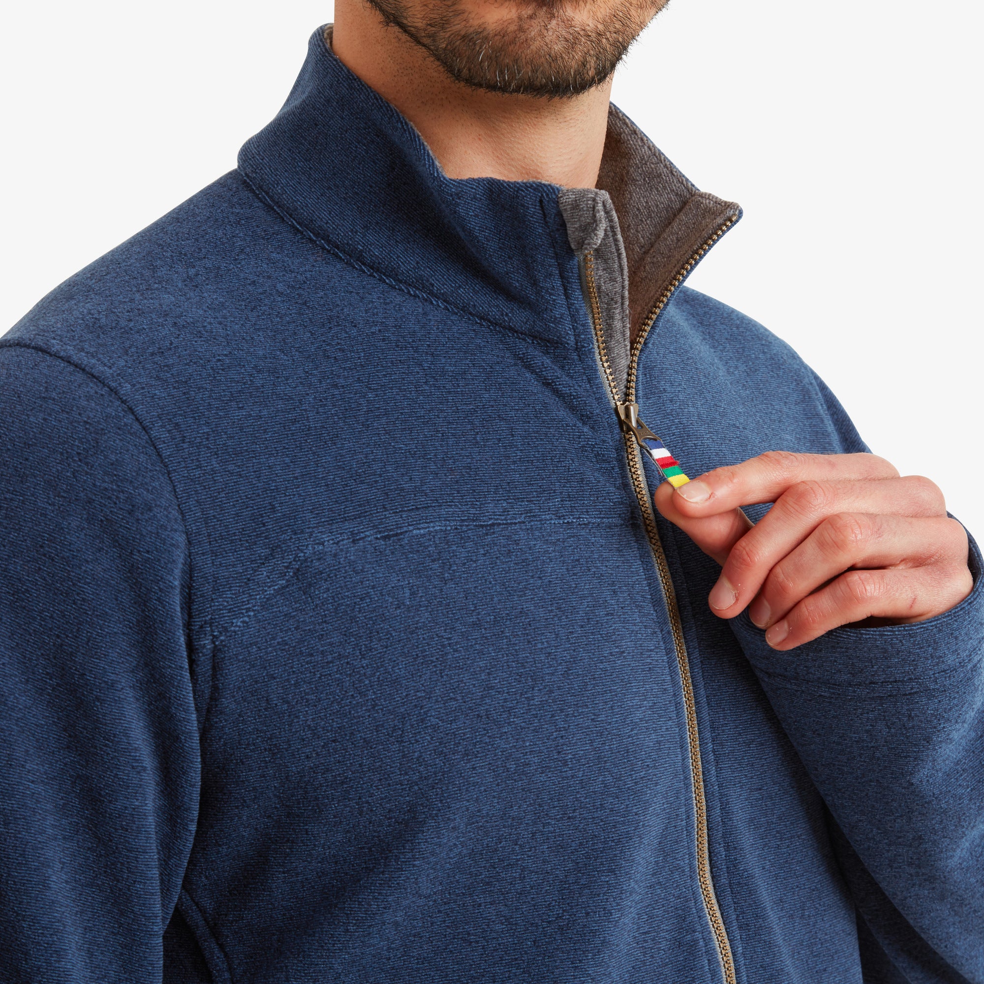 A close-up shot focuses on the zipper, showing a fabric pull tab with colorful stripes, a signature Sherpa design. The soft inner lining is slightly visible, emphasising the comfort of the Sherpa Adventure Gear Rolpa Eco Jacket in Blue’s material.