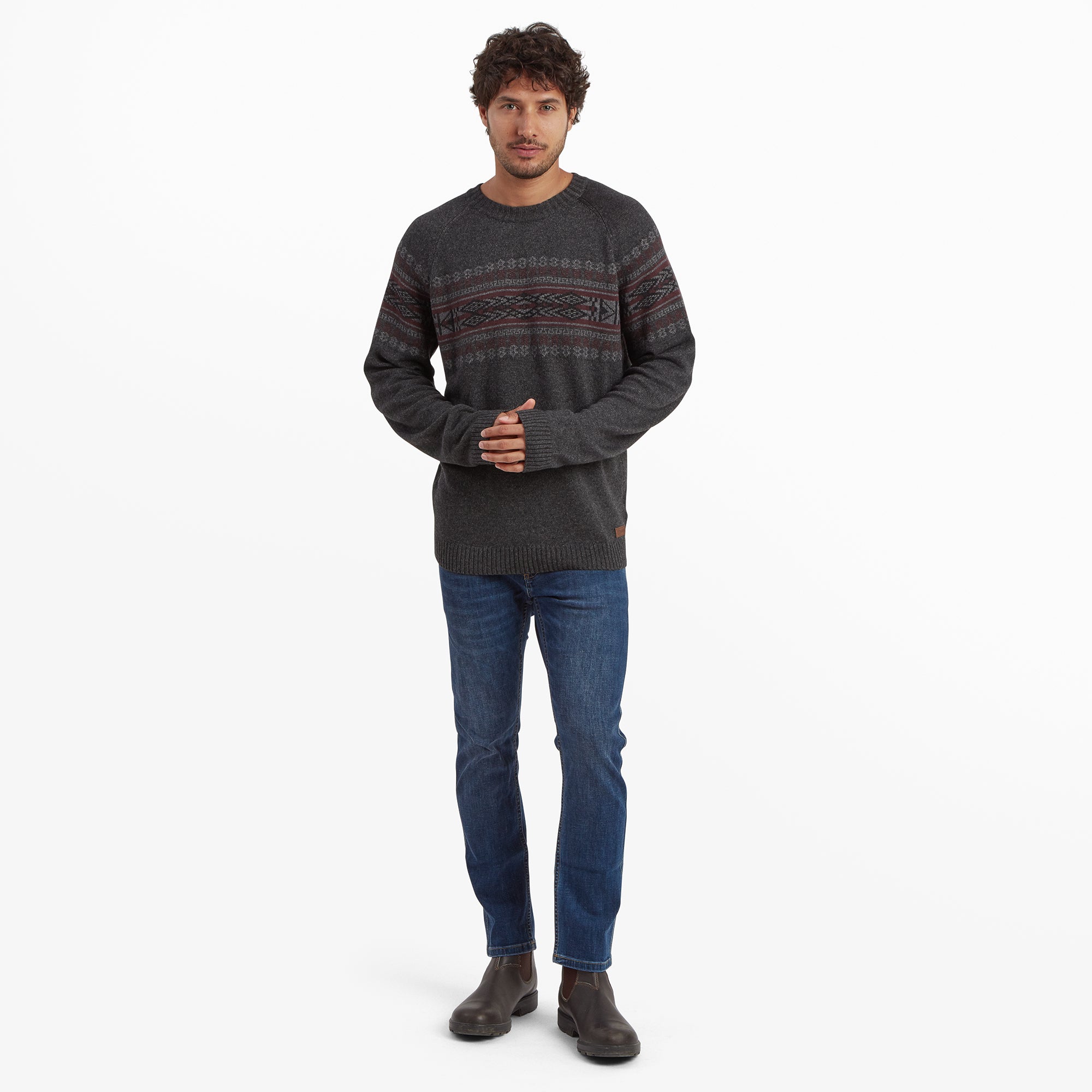 A full-body view of the man wearing the same Sherpa Adventure Gear Sajit Eco Crew in Grey paired with blue jeans and brown boots. He is standing upright, clasping his hands together, highlighting the sweater's length and fit.