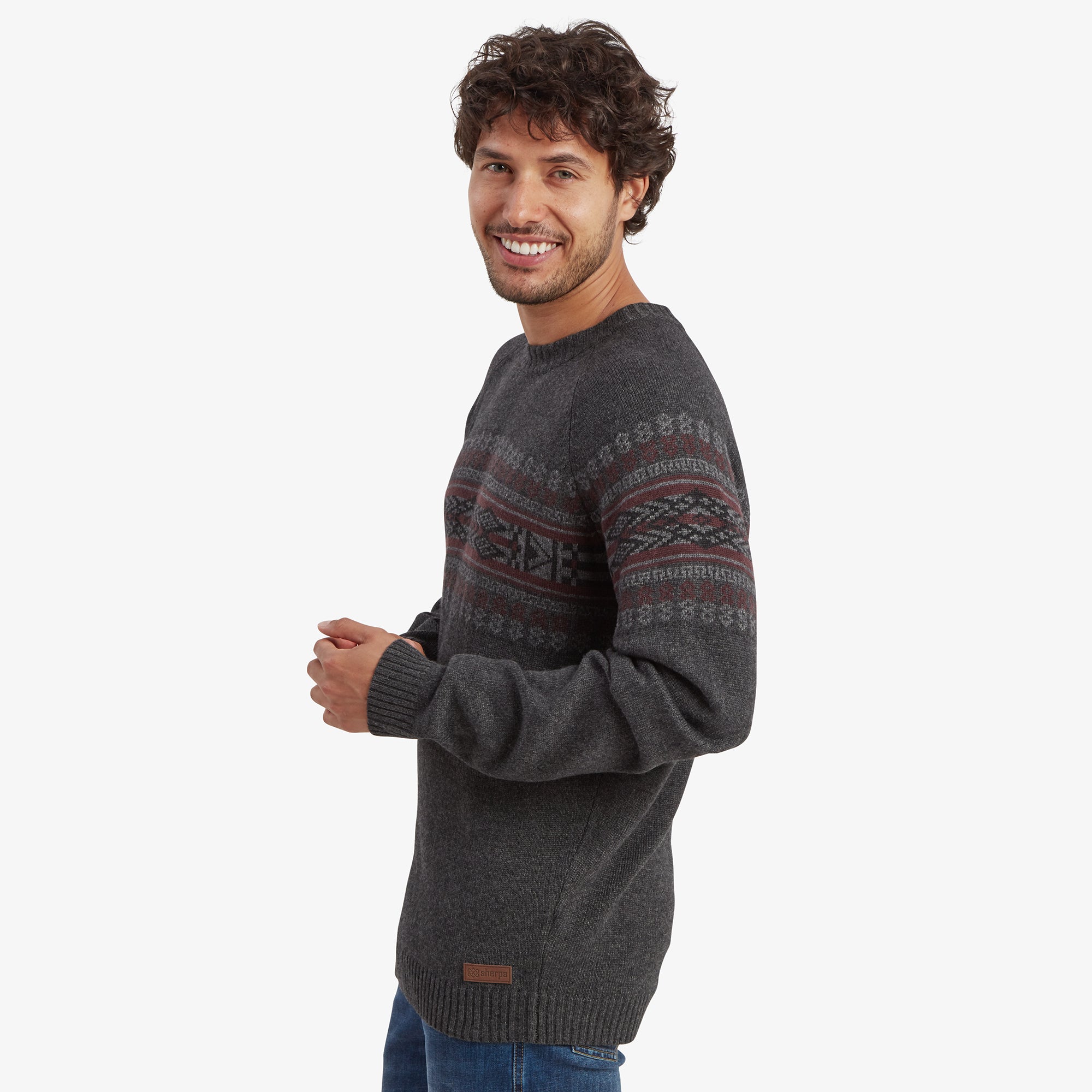 A side profile view of the man in the Sherpa Adventure Gear Sajit Eco Crew in Grey. He is slightly turned, smiling, with one hand casually resting, providing a detailed look at the pattern along the sleeve and a small leather Sherpa logo patch near the hem.