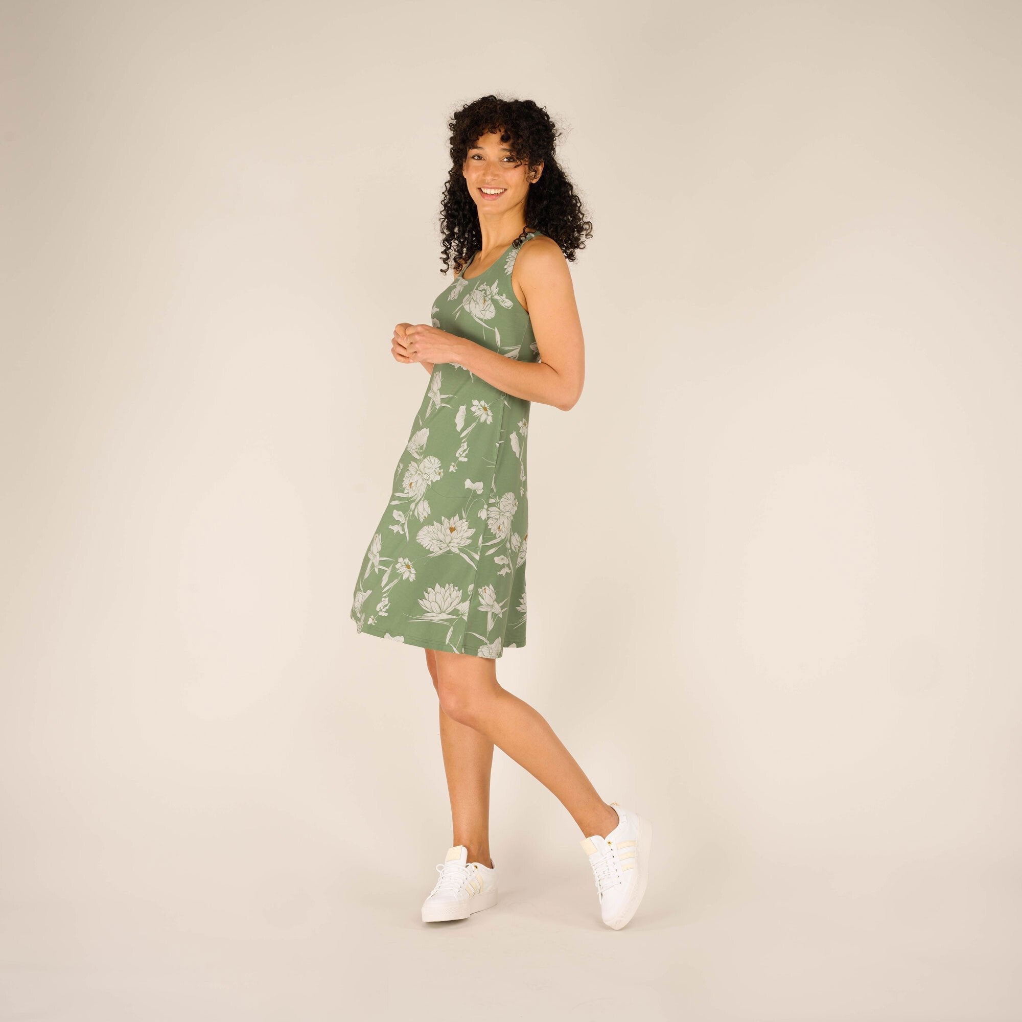 A side view of the woman in the Sherpa Adventure Gear Neha Dress in Green, smiling while slightly turning her upper body. The dress has a flattering silhouette, highlighting her waist. She wears white sneakers, adding a casual touch to the outfit.