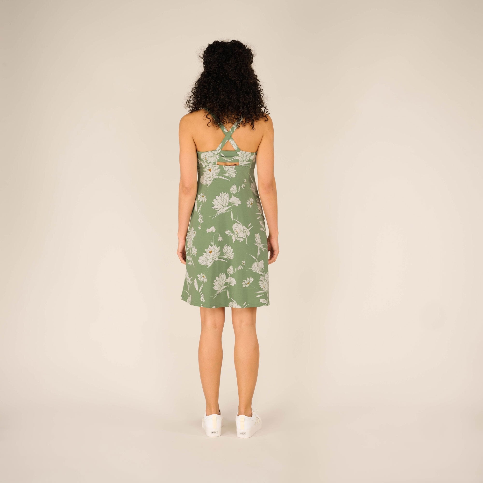 The back view of the Sherpa Adventure Gear Neha Dress in Green reveals a crisscross strap design with a small cutout below the straps. The dress flows just above the knees, and the model stands with her feet slightly apart against a beige background.