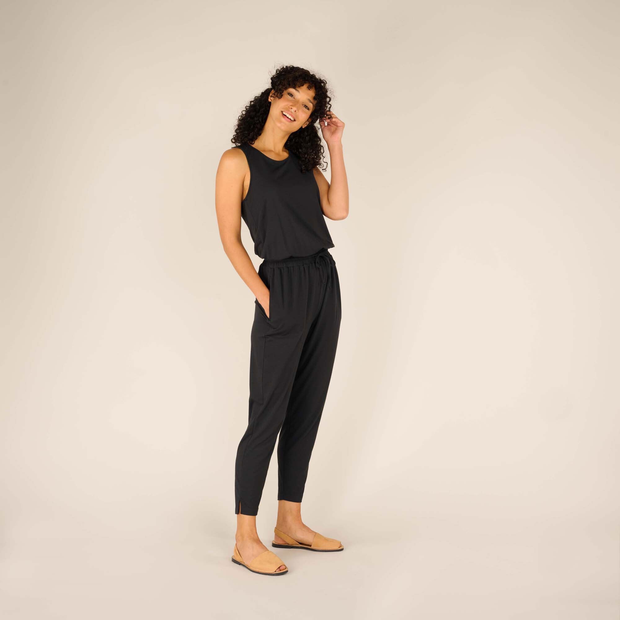 The woman is slightly turned, showcasing the side view of the Sherpa Adventure Gear Neha Jumpsuit in Black. The outfit's drawstring waist and tapered pant legs are visible, emphasising its tailored yet comfortable design. Her hand rests casually in her pocket, and her expression is cheerful.