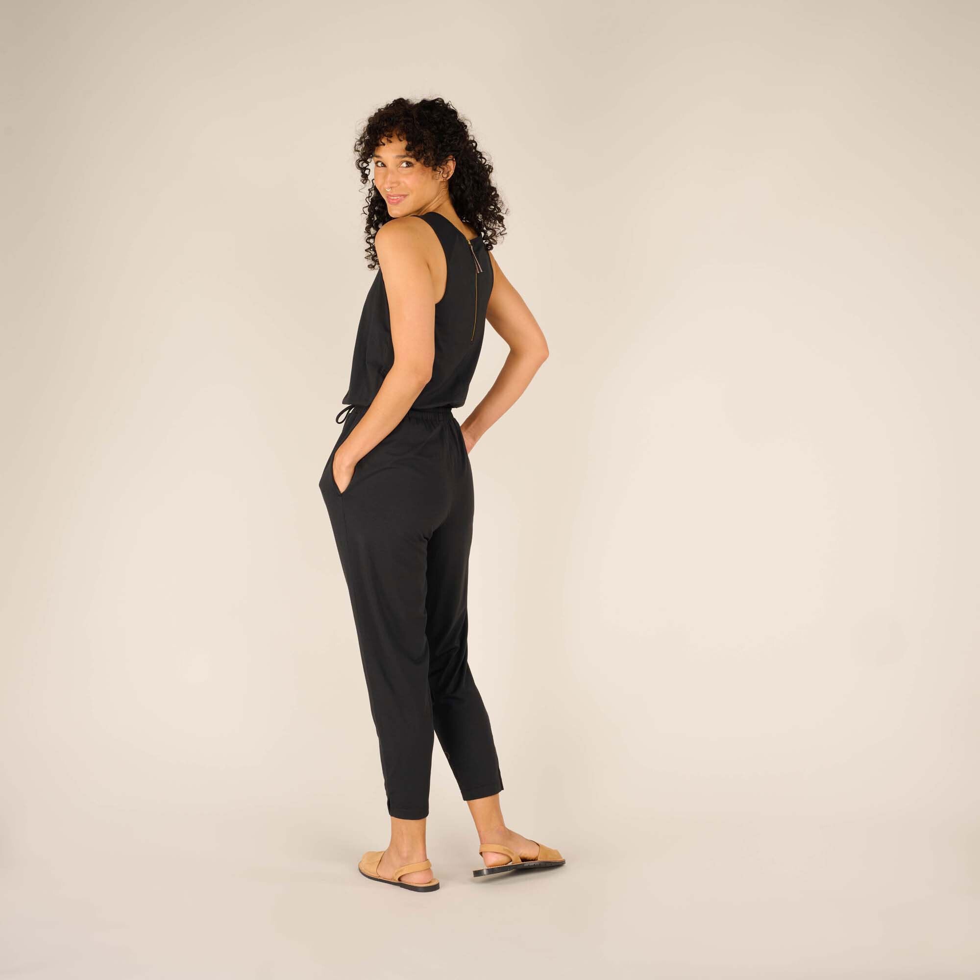 The woman turns slightly backward, giving a view of the back zipper and the relaxed fit of the Sherpa Adventure Gear Neha Jumpsuit in Black’s upper section. Her posture accentuates the comfortable and stylish silhouette of the outfit.