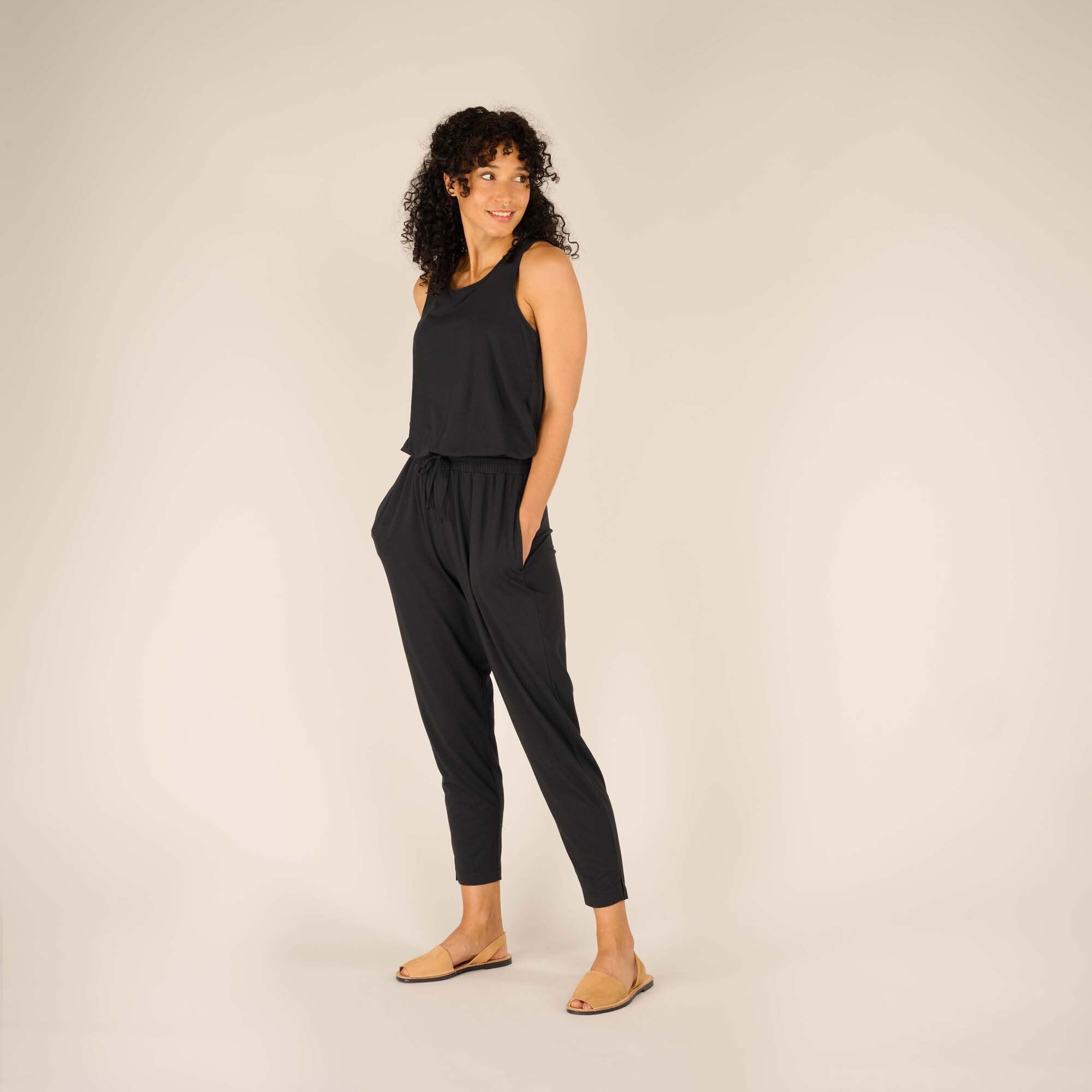 The woman stands in a casual pose with a slight smile, her hands tucked into the Sherpa Adventure Gear Neha Jumpsuit in Black’s side pockets. The neutral beige background enhances the simplicity and elegance of the black jumpsuit.