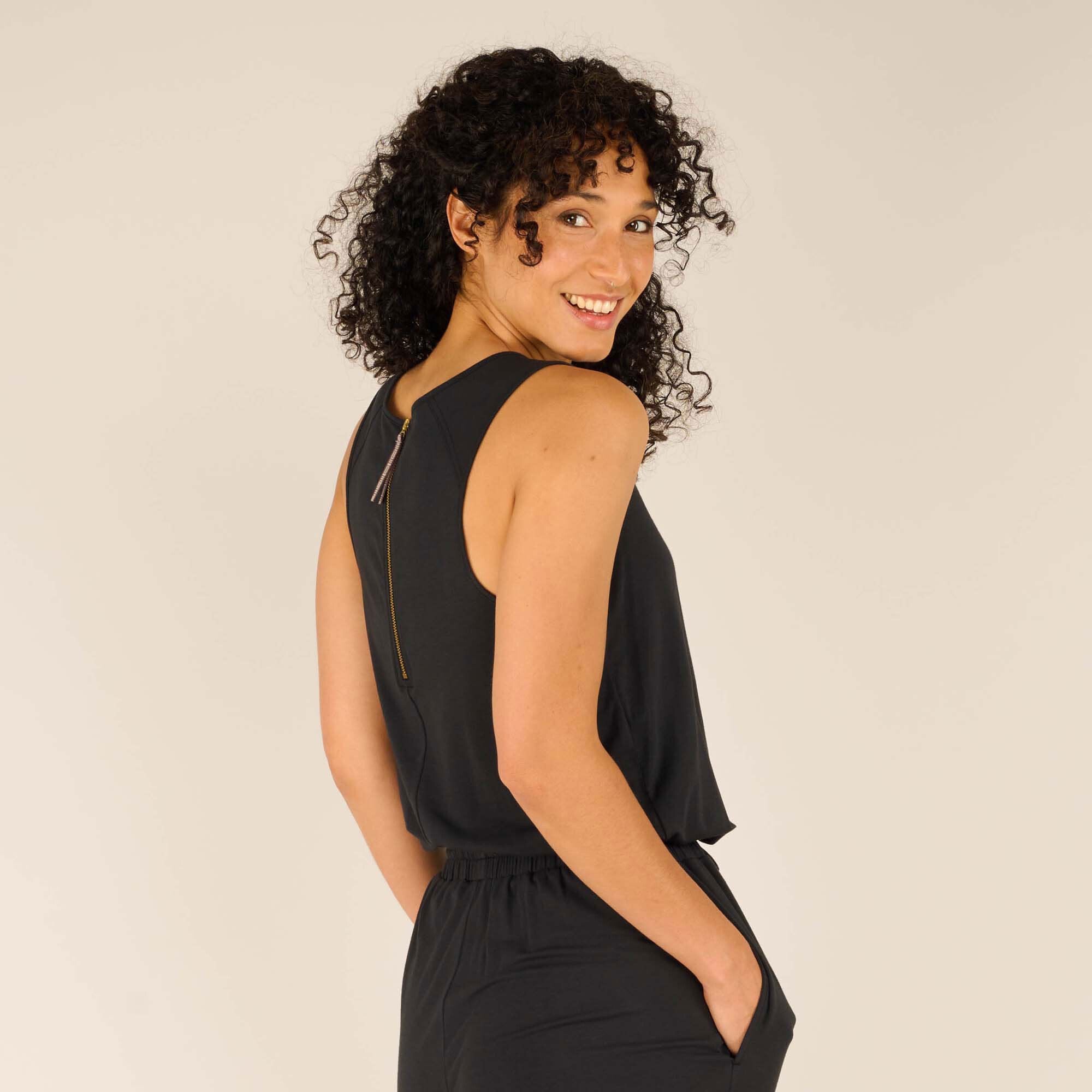 A zoomed-in image showcasing the back of the Sherpa Adventure Gear Neha Jumpsuit in Black, focusing on the gold zipper detail and the smooth, tailored finish of the sleeveless design. The woman smiles warmly over her shoulder, highlighting her curly hairstyle and the garment's elegant construction.