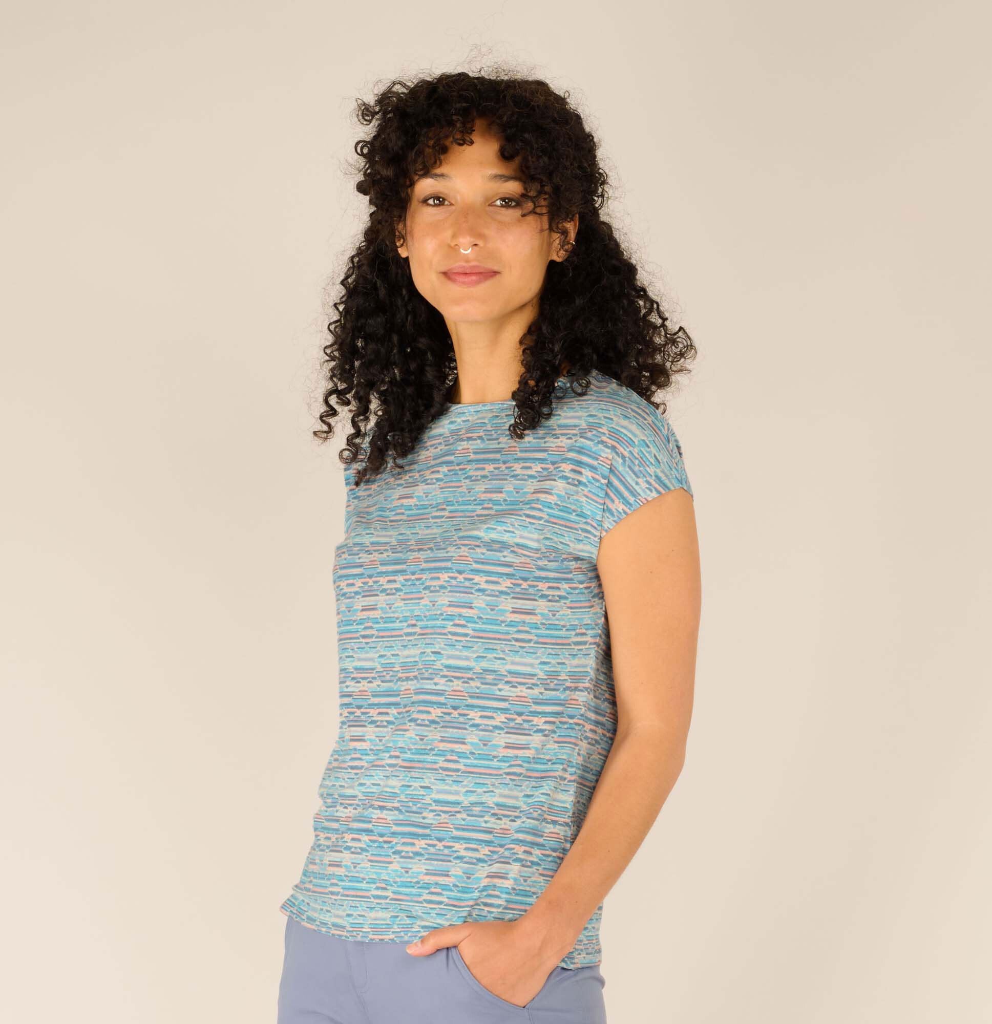 The same woman poses slightly turned to her right, offering a better view of the Sherpa Adventure Gear Neha T-Shirt in Blue’s pattern. Her soft smile and confident stance highlight the shirt’s comfortable fit. She has her left hand casually tucked into her pocket.