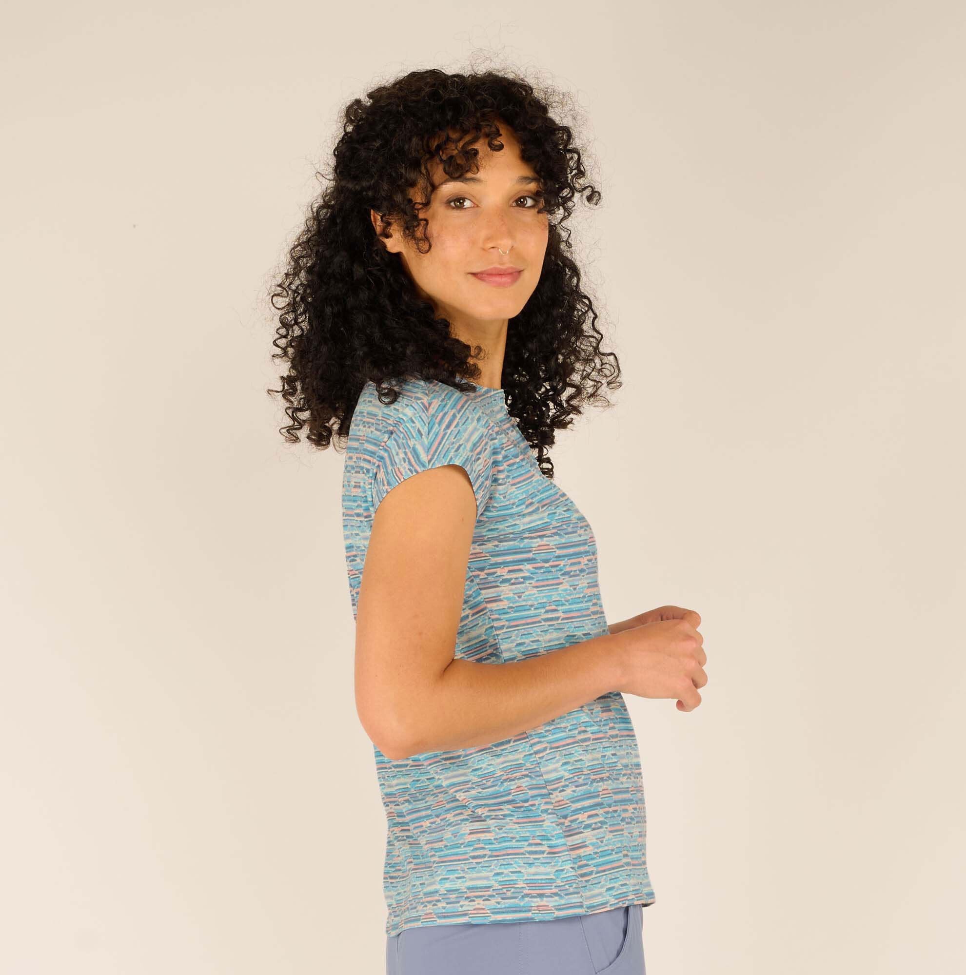 A side profile of the woman, showcasing the Sherpa Adventure Gear Neha T-Shirt in Blue’s sleek fit against her frame. The cap sleeves and round neckline are visible, with her curly hair falling naturally over her shoulder.