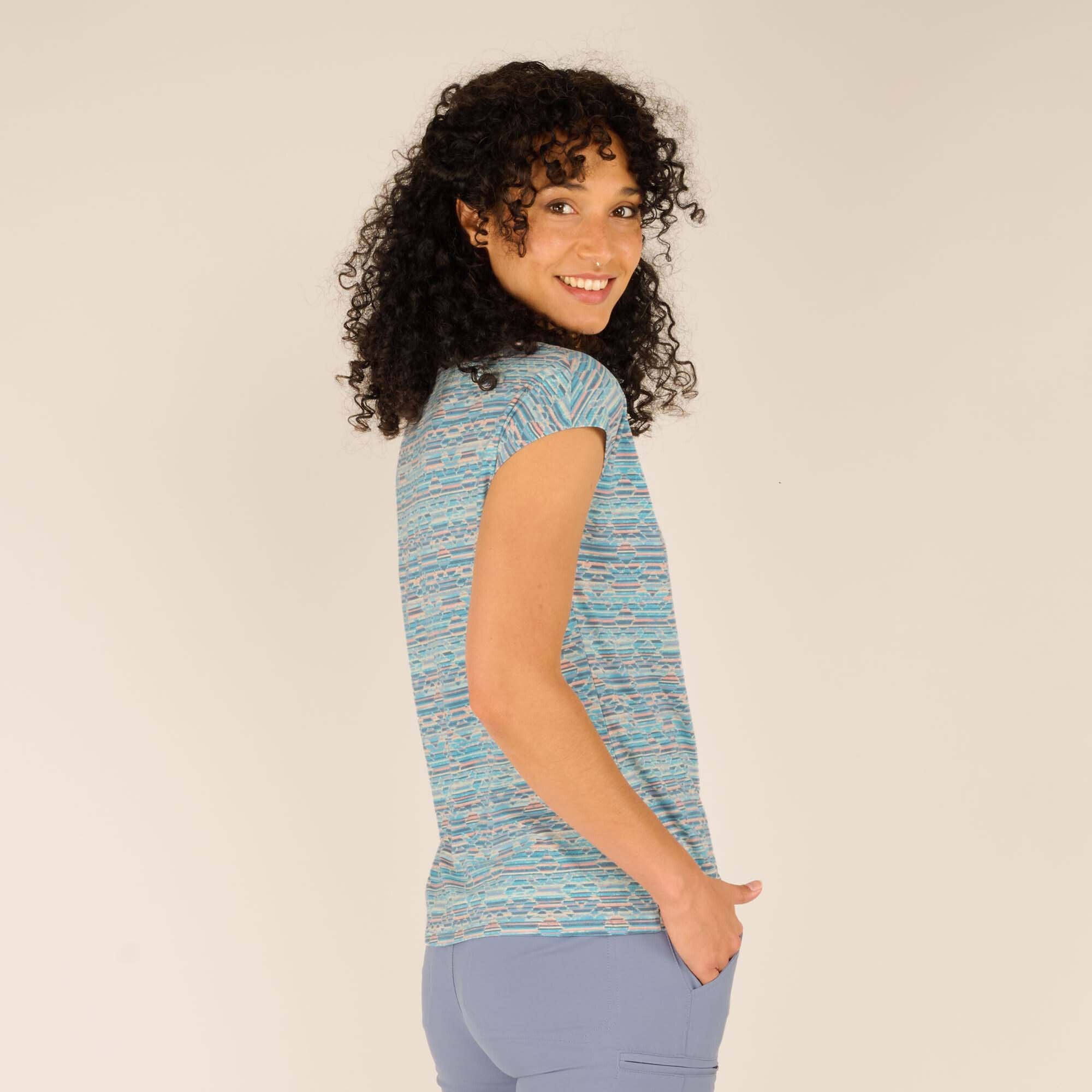 A three-quarter rear view of the model, where she turns her head slightly to smile at the camera. This perspective highlights the Sherpa Adventure Gear Neha T-Shirt in Blue’s structure and how it contours to her body. She wears light blue casual pants, complementing the t-shirt’s color scheme.